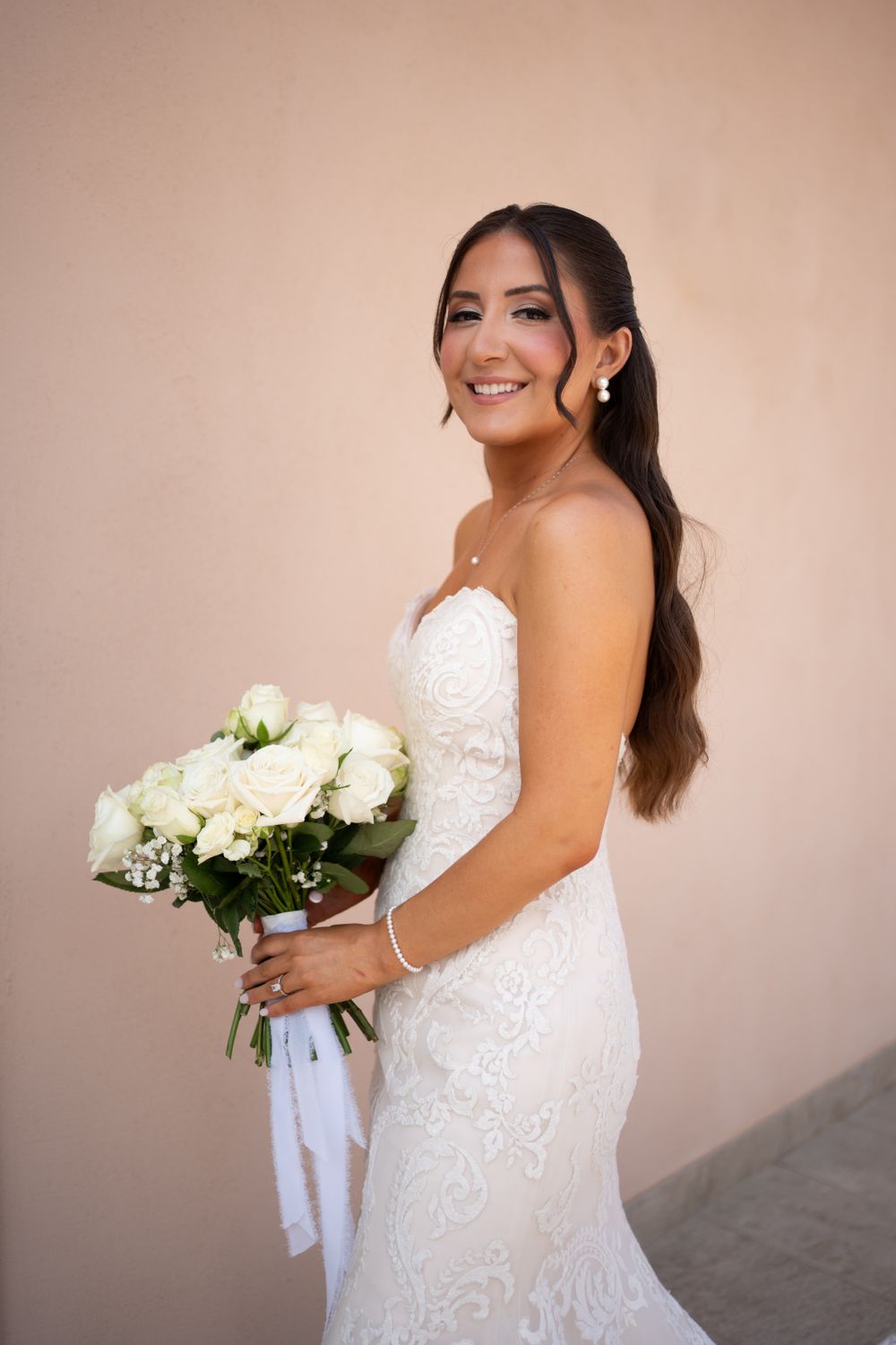 Bride in fitted lace gown smiles while holding white rose bouquet.