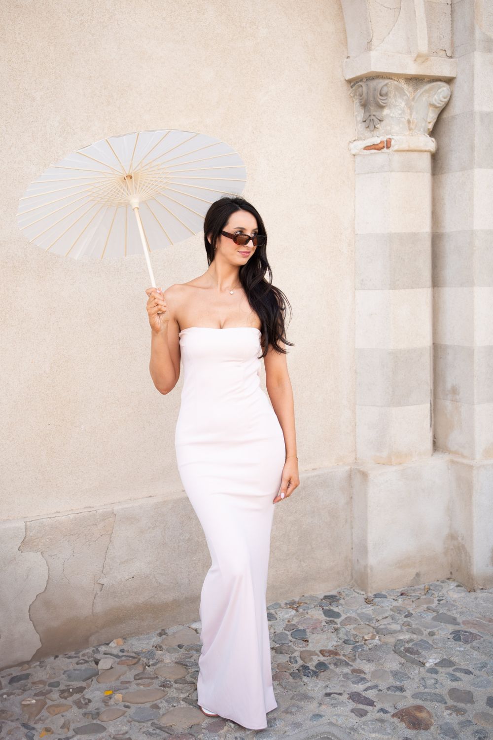 A figure in a white dress poses with a parasol against a textured stone wall.