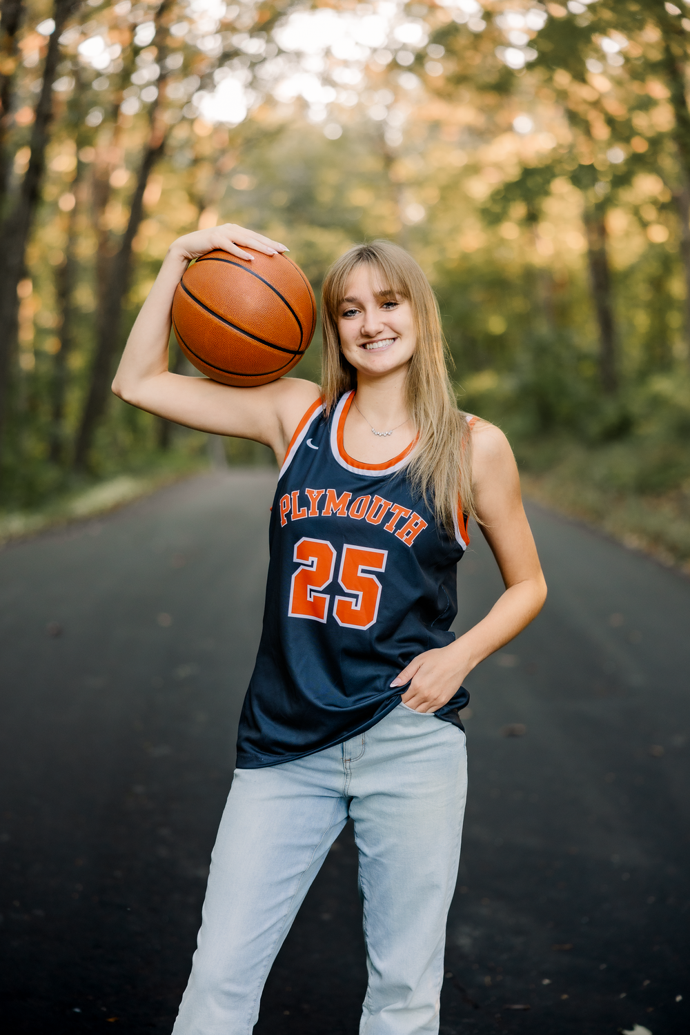 Basketball player in navy blue jersey number 23 and light jeans poses playfully with ball in outdoor setting.