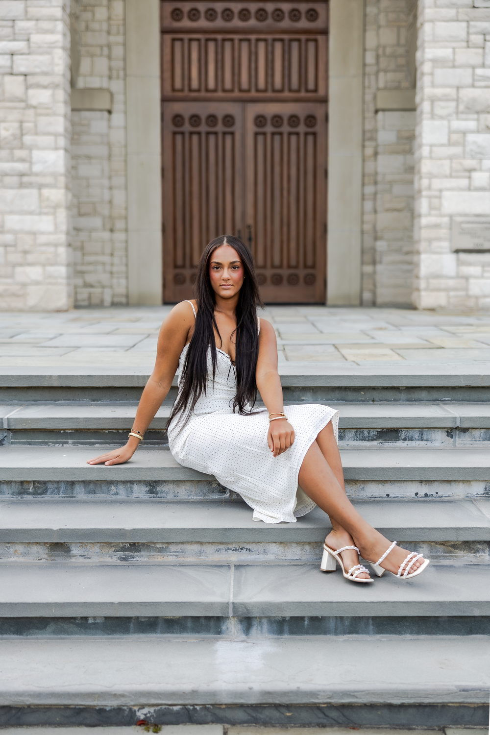 A person in a white dress sits gracefully on concrete steps outside a building with ornate wooden doors.