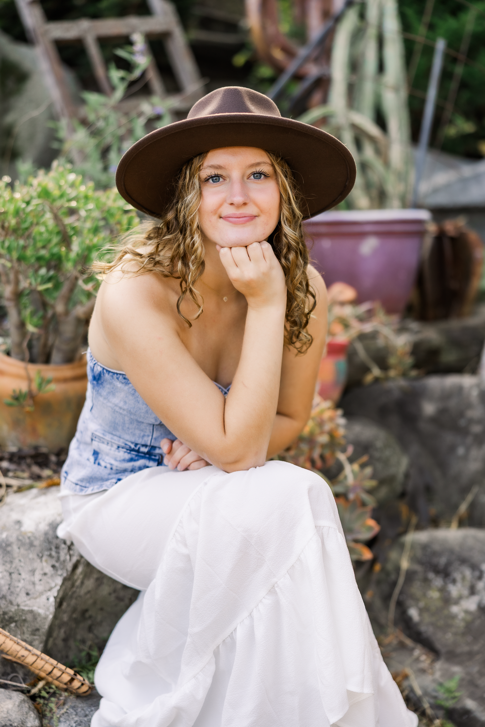 A person in a wide-brimmed brown hat and white dress poses outdoors among garden plants and stone features.