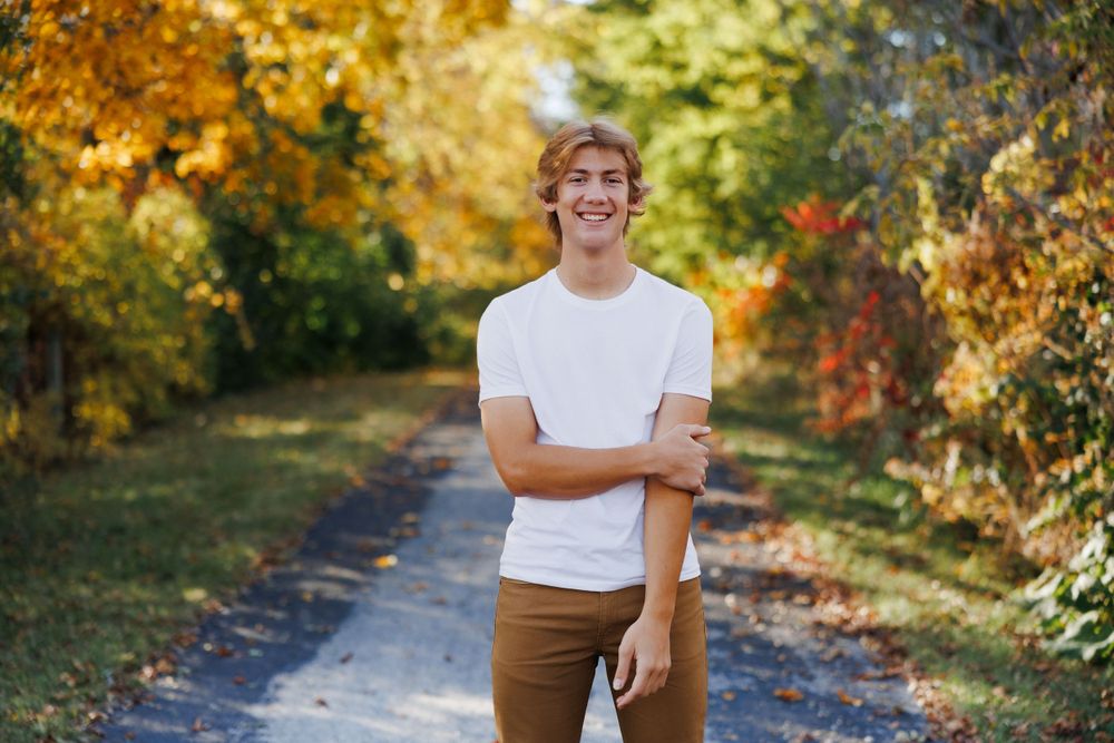 High school senior boy standing on a pathway surrounded by vibrant fall foliage.