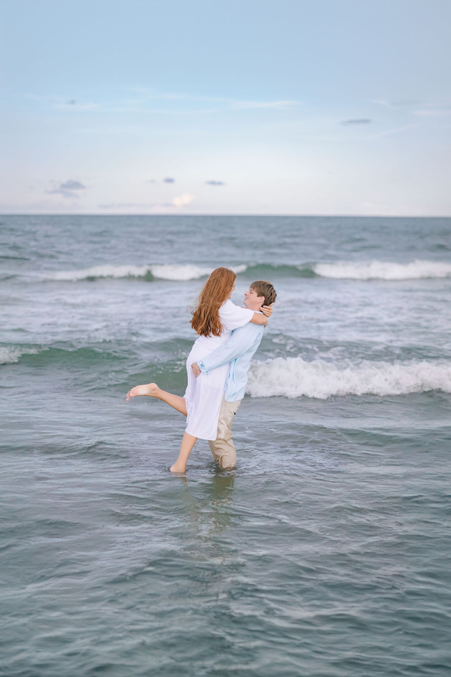 Couple embraces while standing in ocean waves during sunset beach photoshoot.