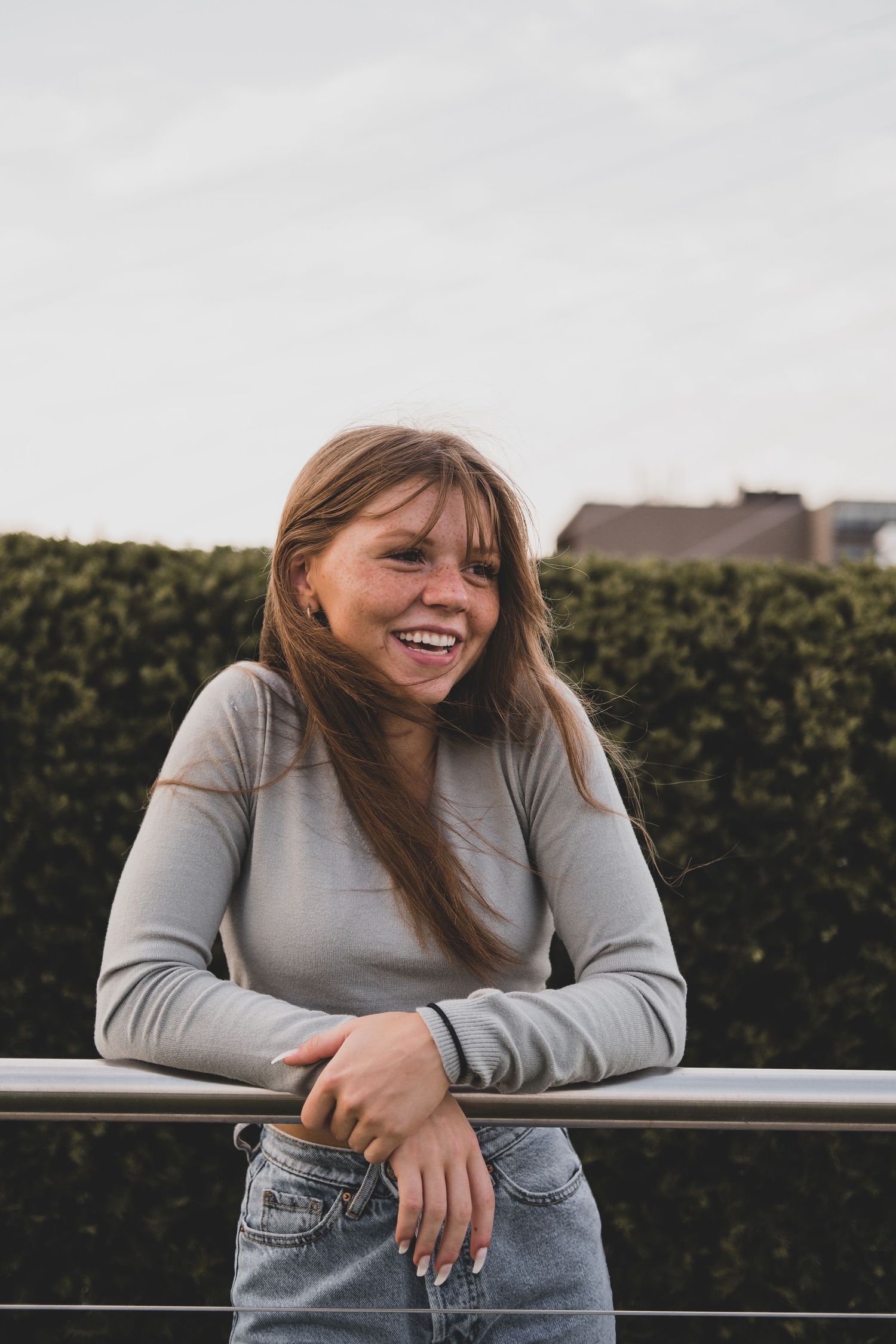 Person in grey sweater leaning on railing while laughing outdoors.