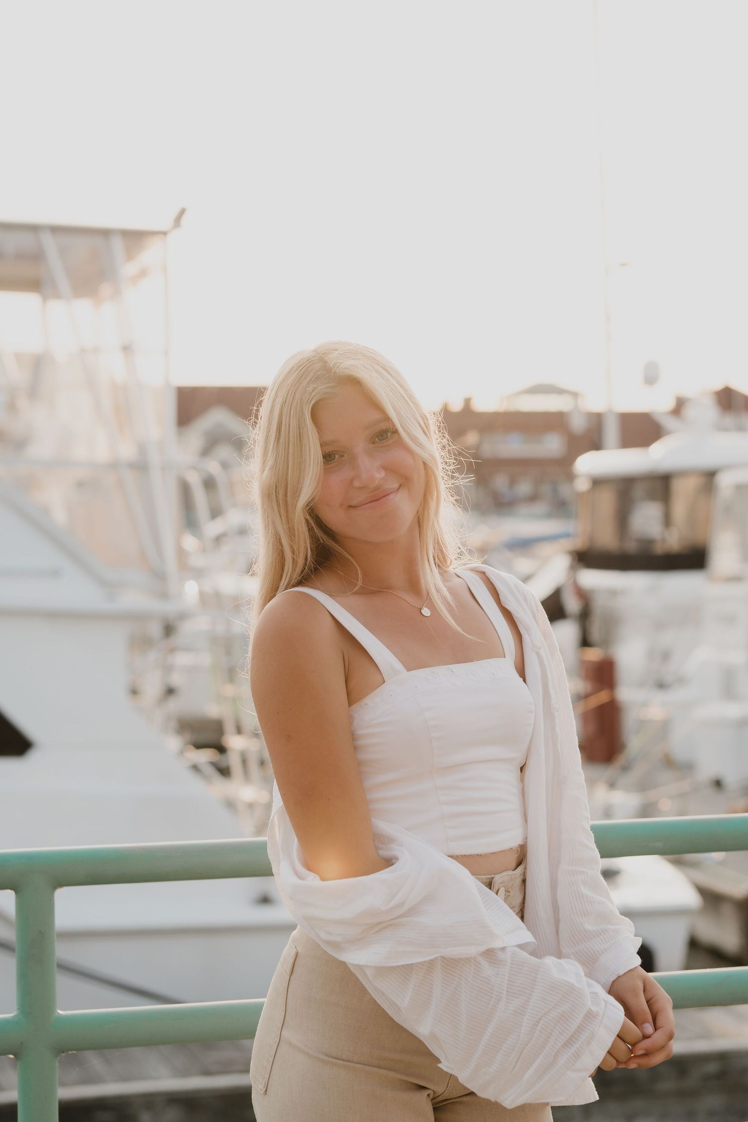 Person in white outfit poses casually on marina deck with boats in background.