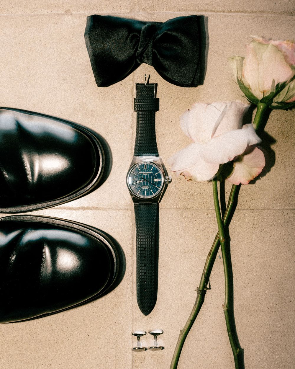 Wedding accessories including dress shoes, bow tie, watch and roses arranged on beige background.