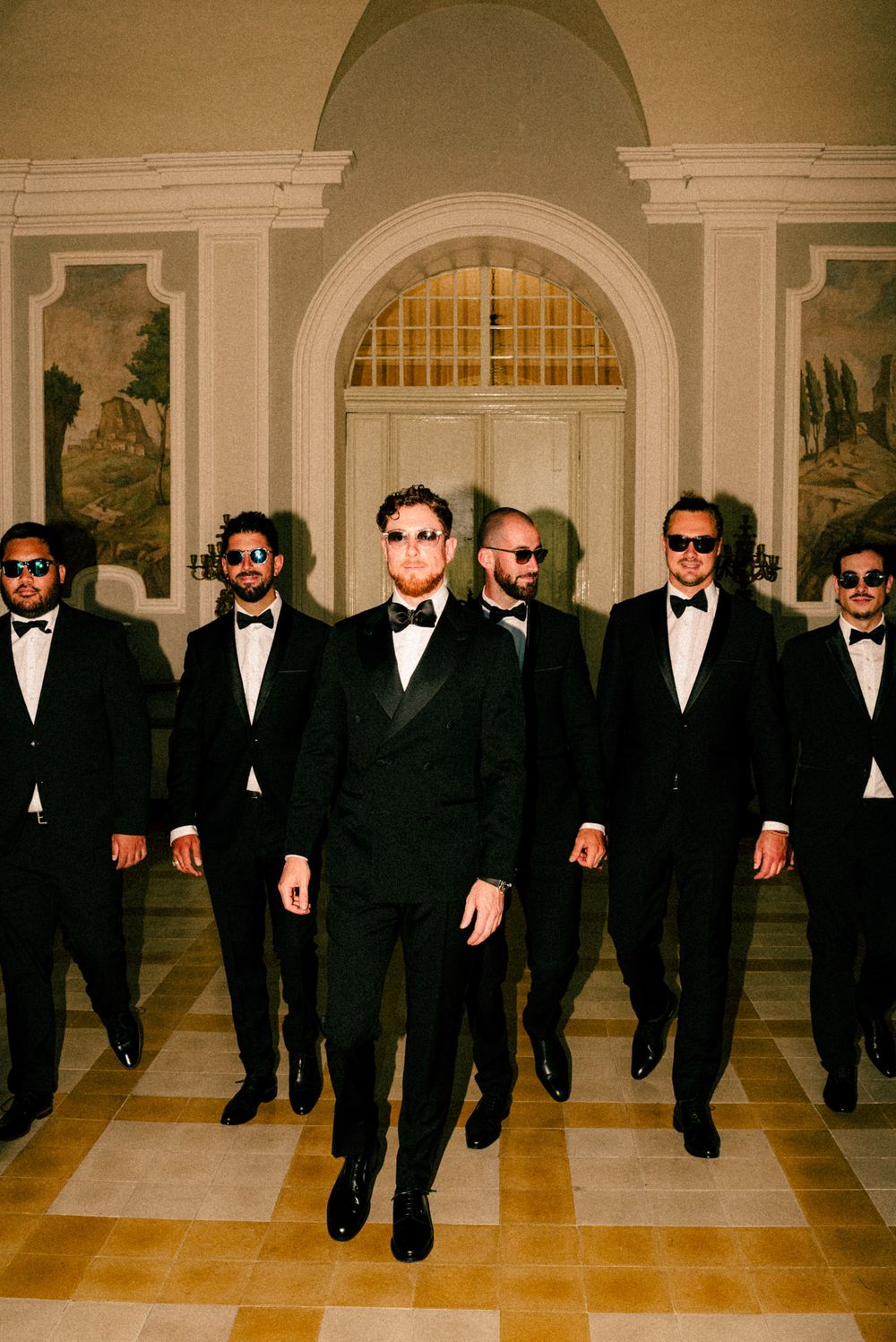 Group of groomsmen in formal black tuxedos and sunglasses walking in formation.