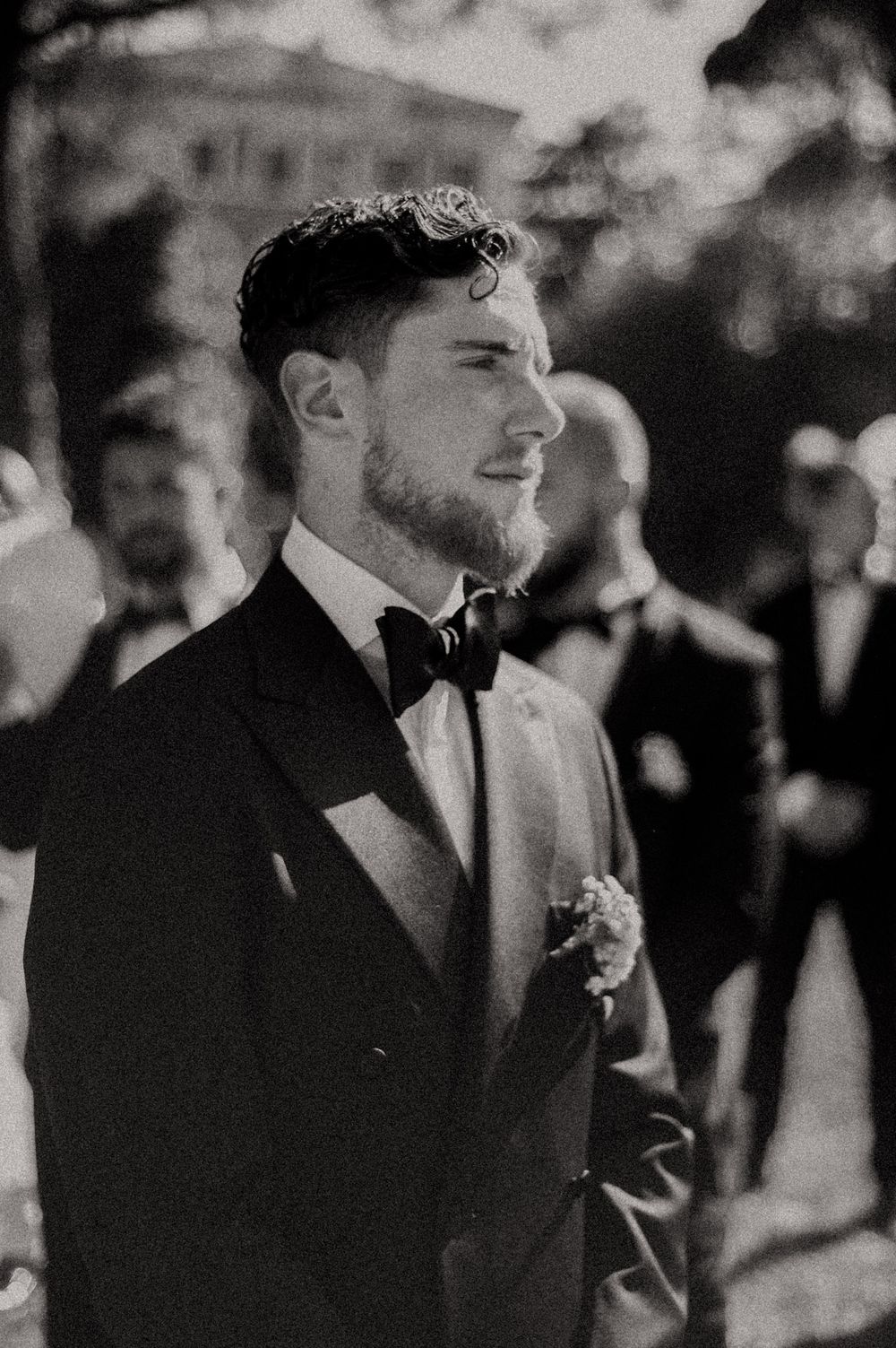 Black and white portrait of someone in a bowtie smiling during an outdoor wedding.