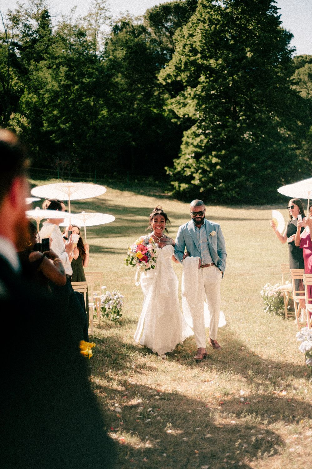 Outdoor wedding ceremony with white parasols and guests watching couple walking down aisle.