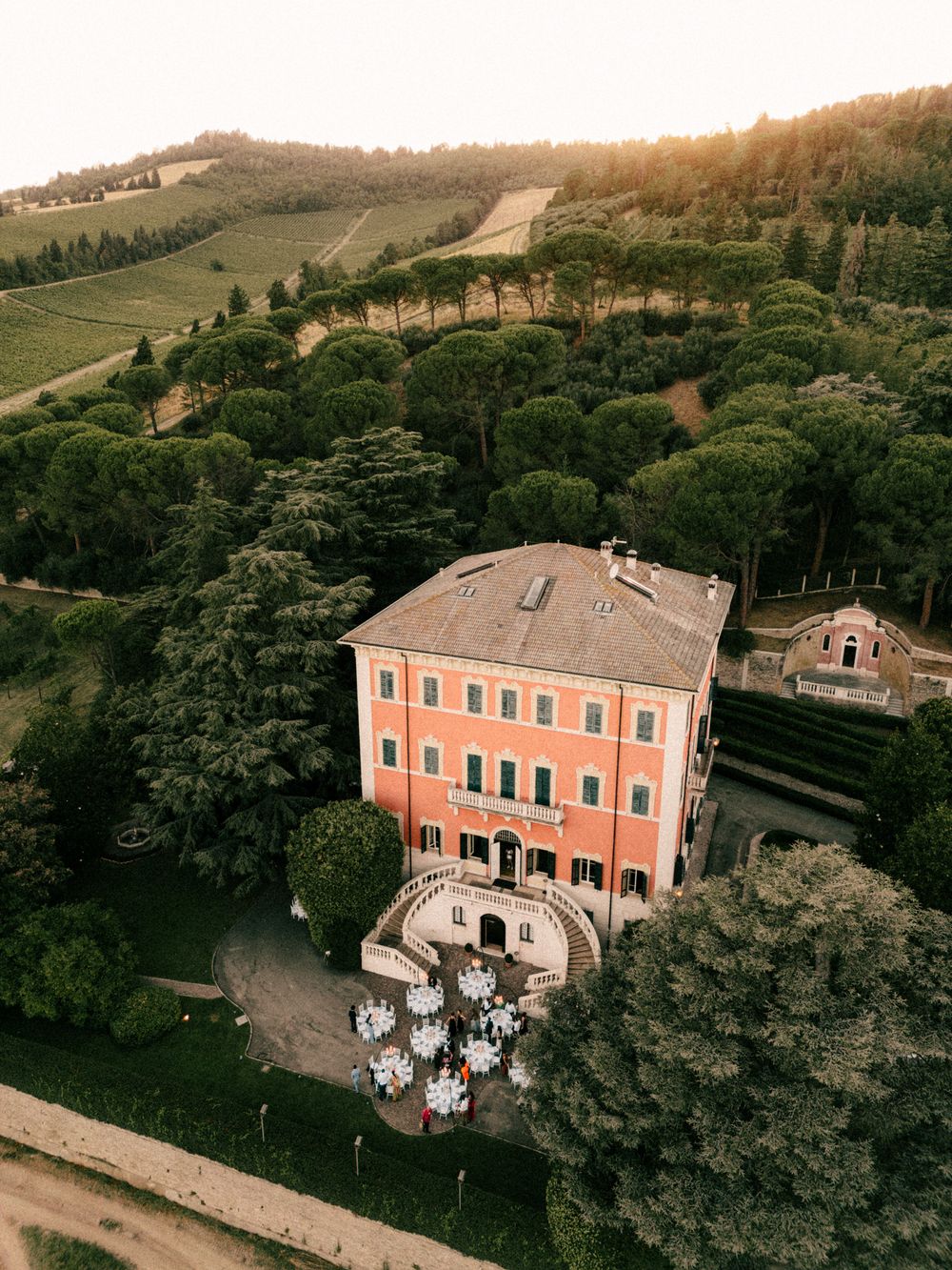Aerial view of a pink Italian villa with manicured gardens and rolling hills in background.