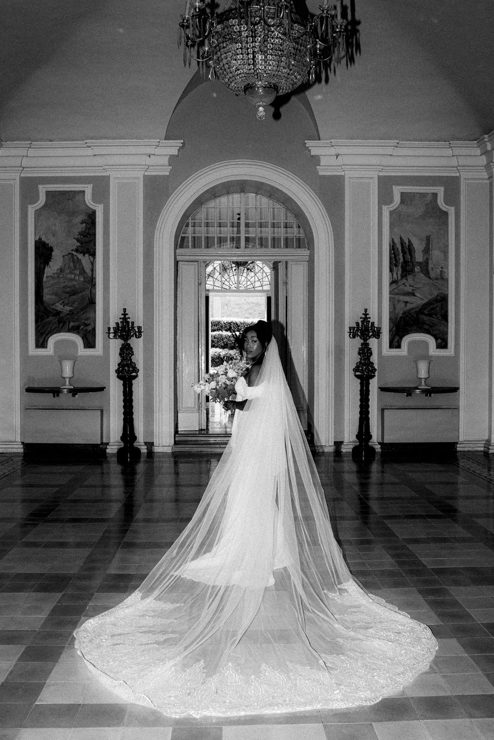Black and white photo of bride in long cathedral veil standing in elegant hallway.
