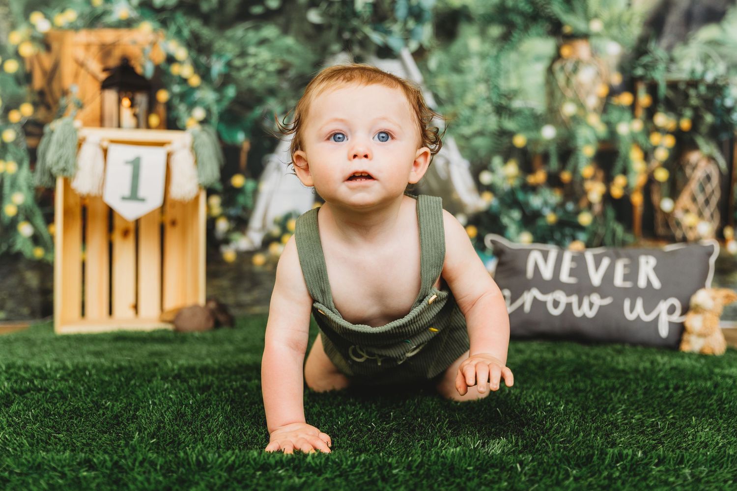 Baby in olive green romper crawling on grass with outdoor party decorations in background.