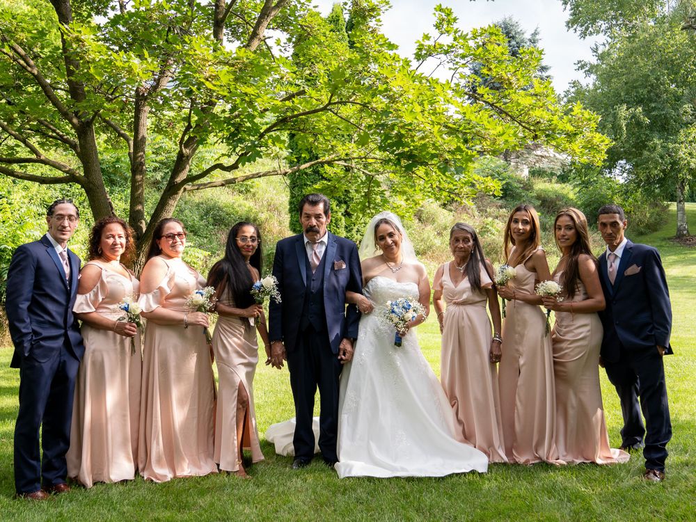 Wedding party with bridesmaids in blush dresses and groomsmen in navy suits pose outdoors under green trees.