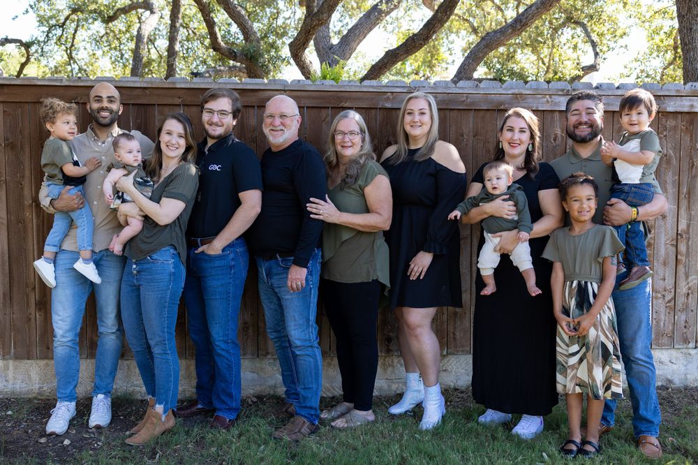 Extended family group photo outdoors with multiple generations posing together against wooden fence.