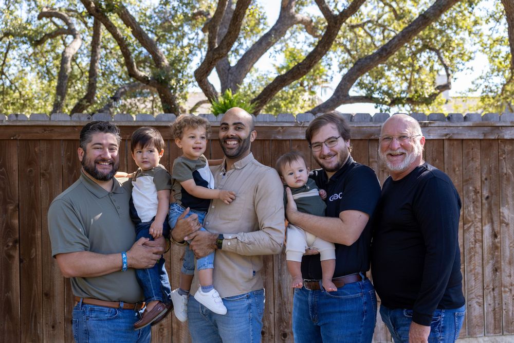 Multi-generational group gathered outdoors for a casual portrait under a large tree against a wooden fence backdrop.