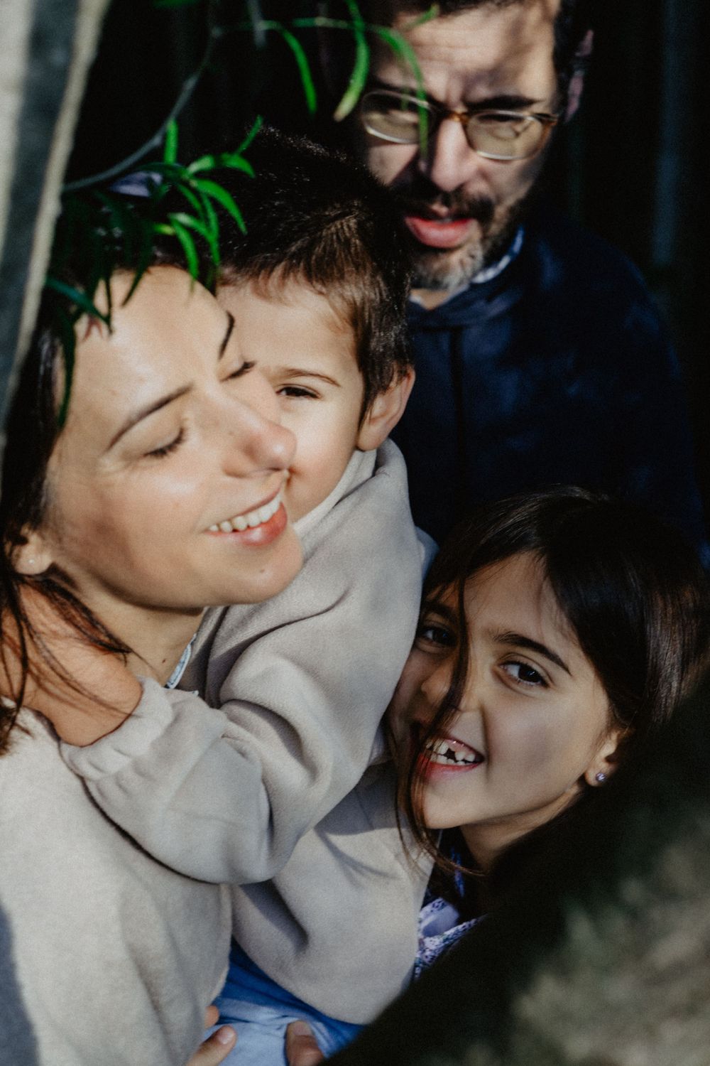 Família abraçada num momento de ternura à sombra da natureza, com sorrisos e olhares cheios de amor e cumplicidade.