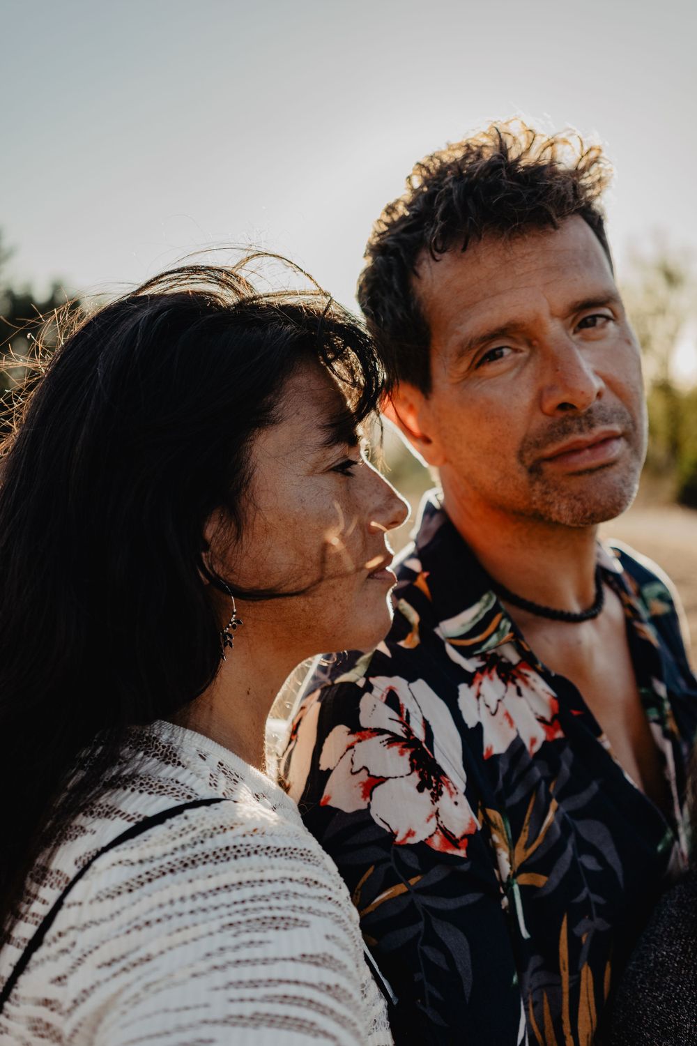 Casal em momento íntimo ao pôr do sol, com expressões intensas e luz dourada destacando detalhes das roupas e rostos ao ar livre.