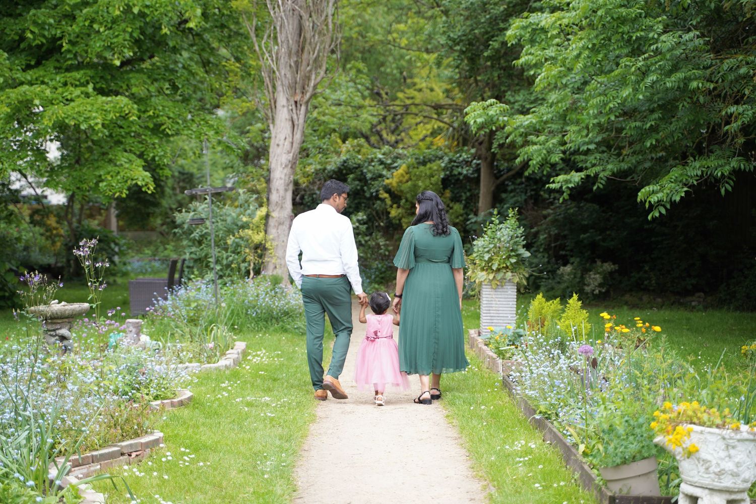 A family photoshoot of the mother and father holding their child's hand and walking down a path lined with purple flowers and lush greenery.