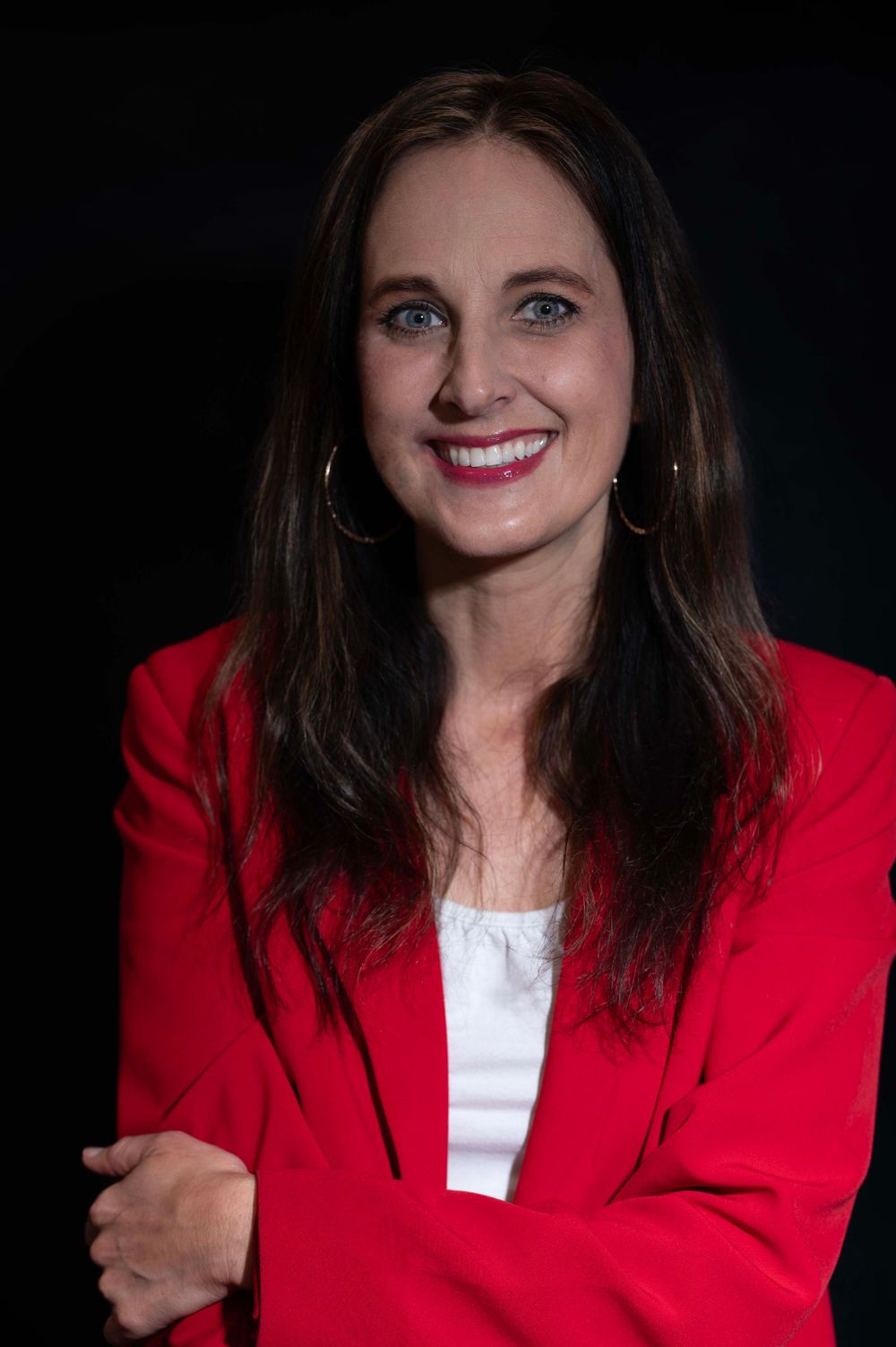 Woman in red blazer with dark hair and blue eyes smiling confidently against dark background.