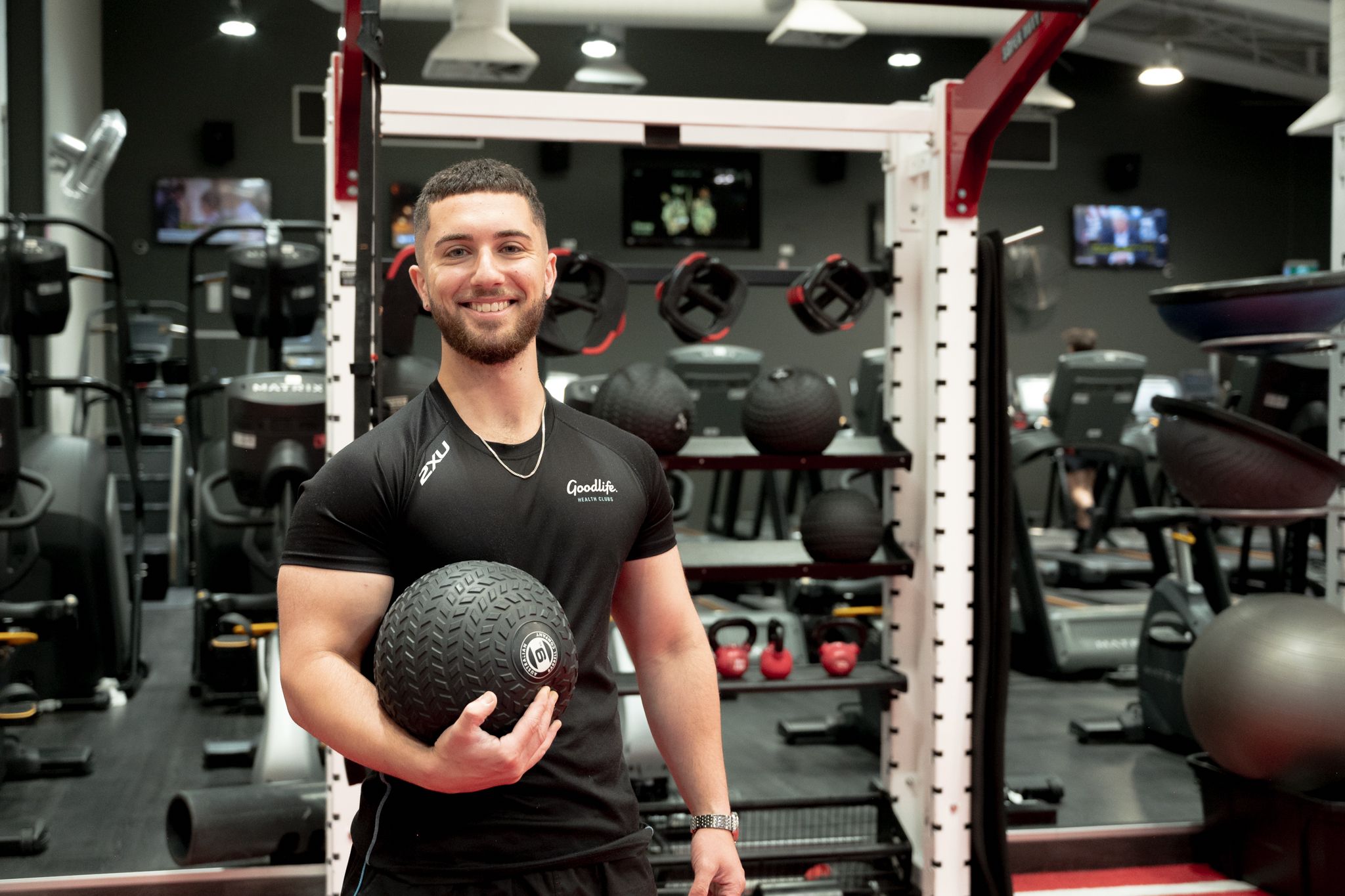 Fit young man smiles holding a medicine ball in a modern gym with equipment in the background.