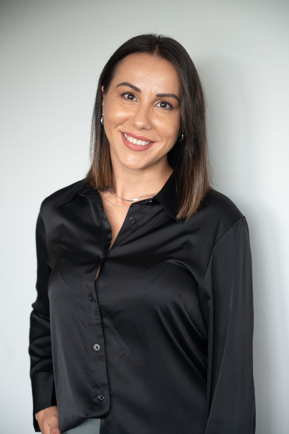 Professional woman with dark hair smiles confidently wearing a black satin button-up blouse against a light background.
