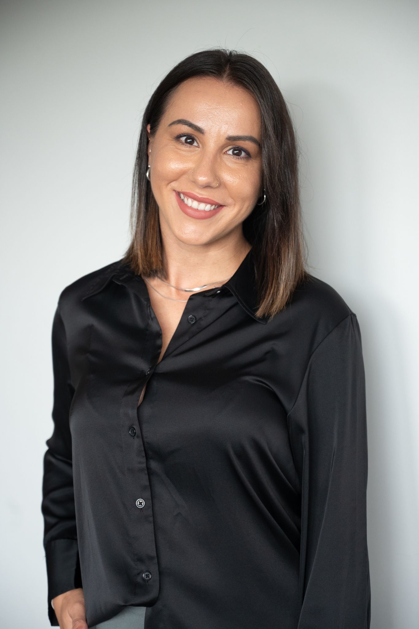 Professional woman with dark hair smiles confidently wearing a black satin button-up blouse against a light background.