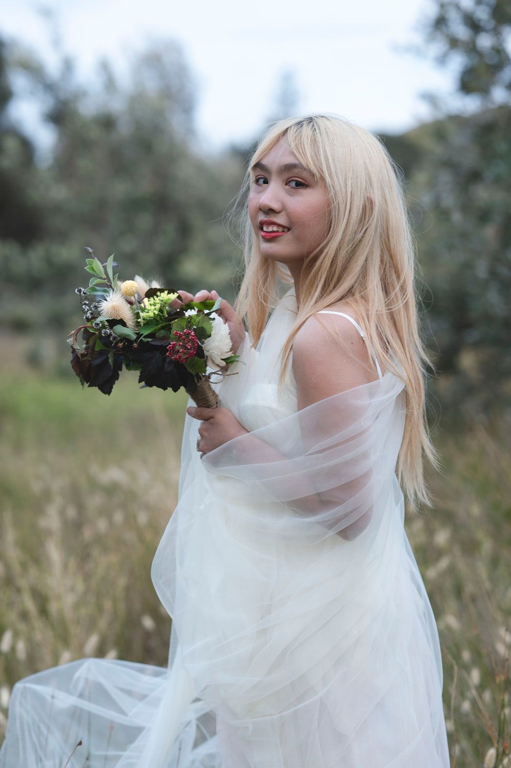 Blonde bride in white tulle gown holding floral bouquet in outdoor field setting.
