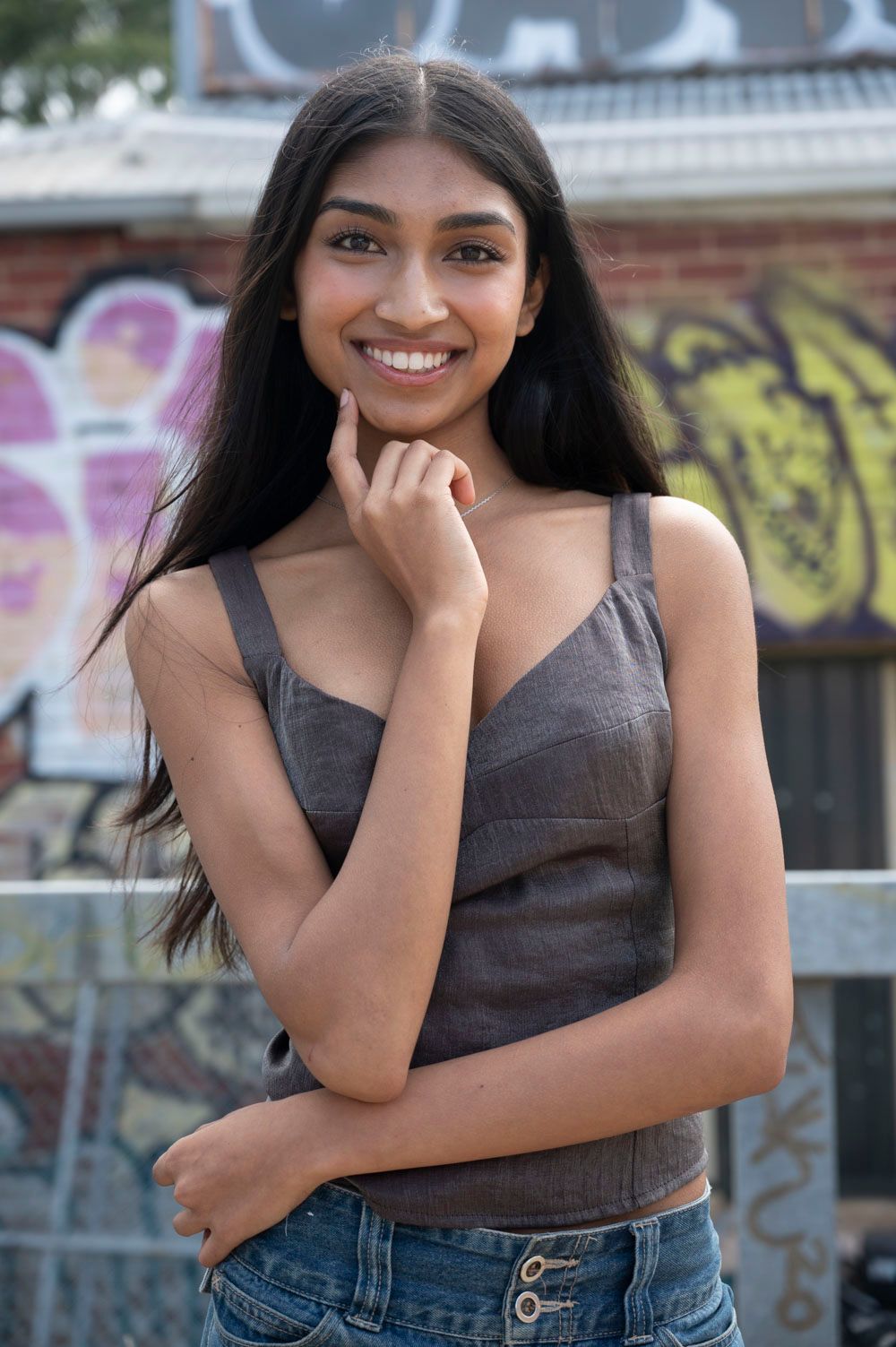 Smiling young woman with long black hair in a gray top and jeans poses near graffiti wall.