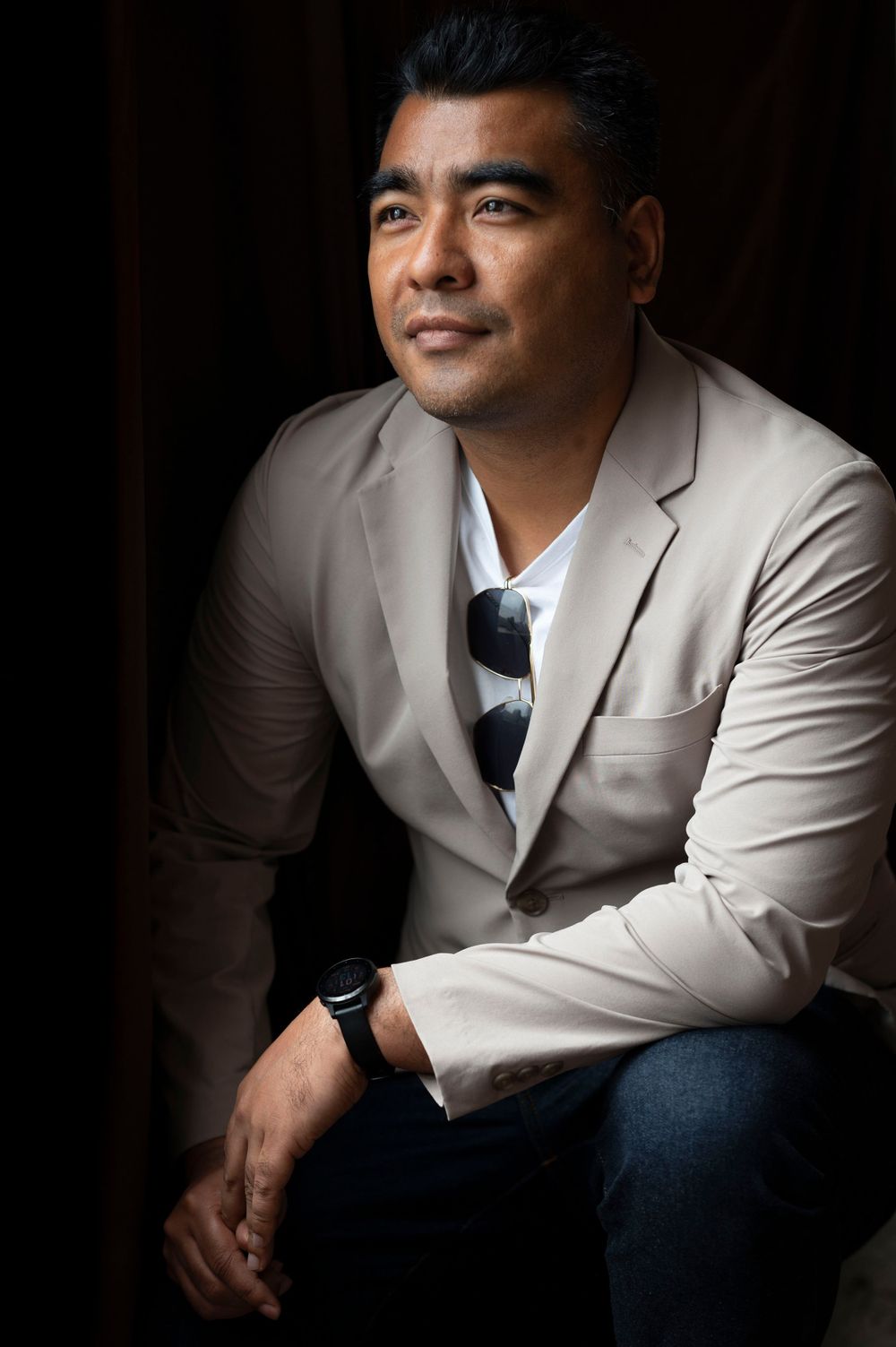 Man in beige blazer sitting thoughtfully with striped tie in dramatic studio lighting.