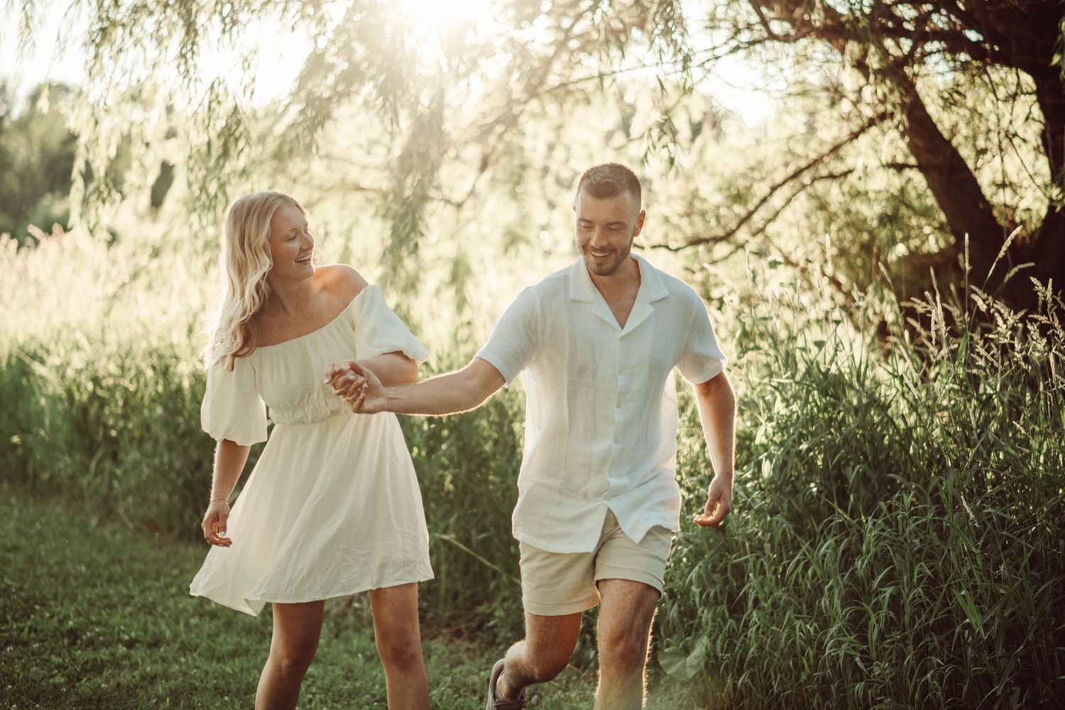 A couple holding hands while running through a golden-lit garden with willow trees.