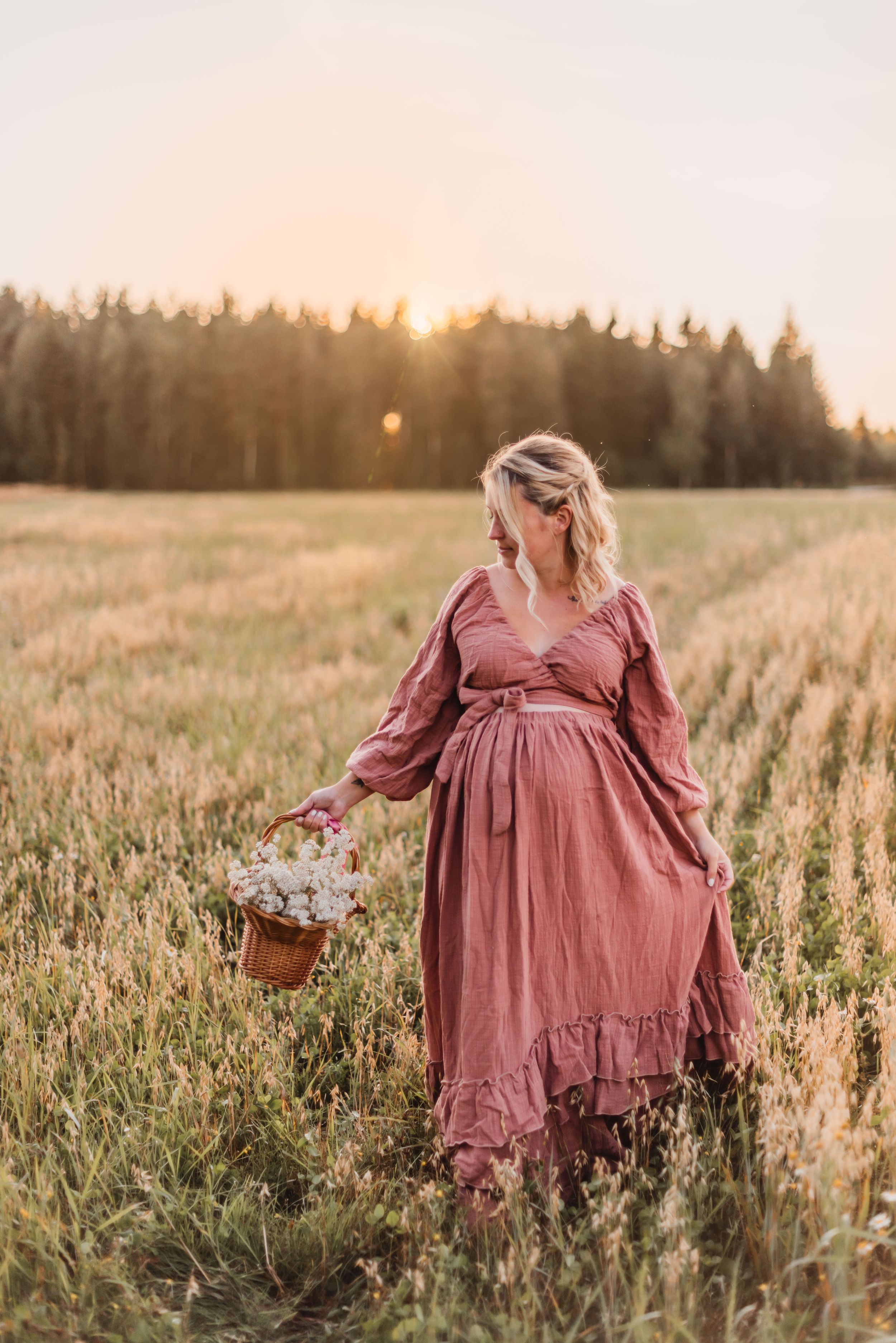A woman in a flowing mauve dress walks through a golden wheat field at sunset, carrying a woven basket.