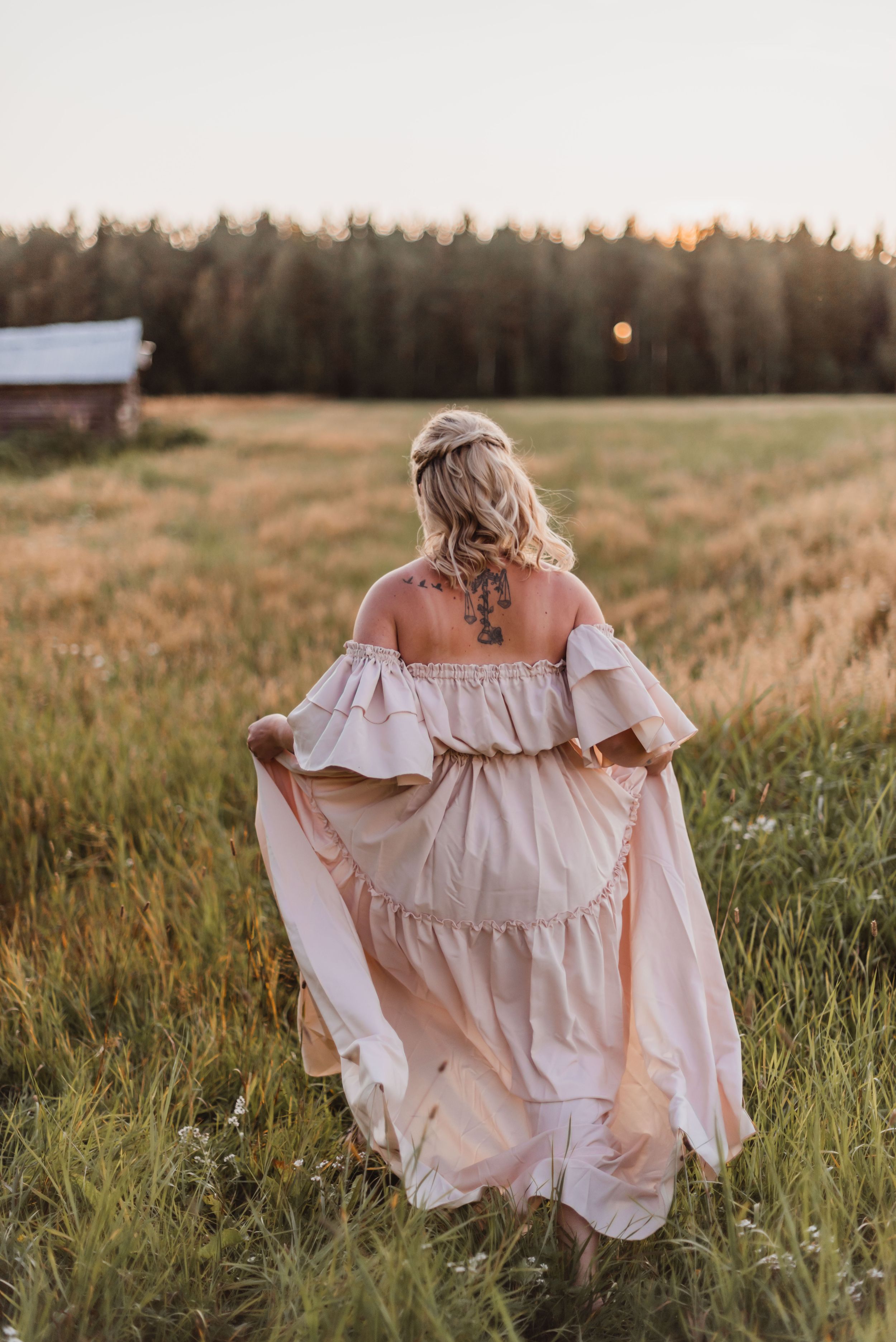 Woman in flowing off-shoulder cream dress walks through golden field at sunset with rustic barn in background.