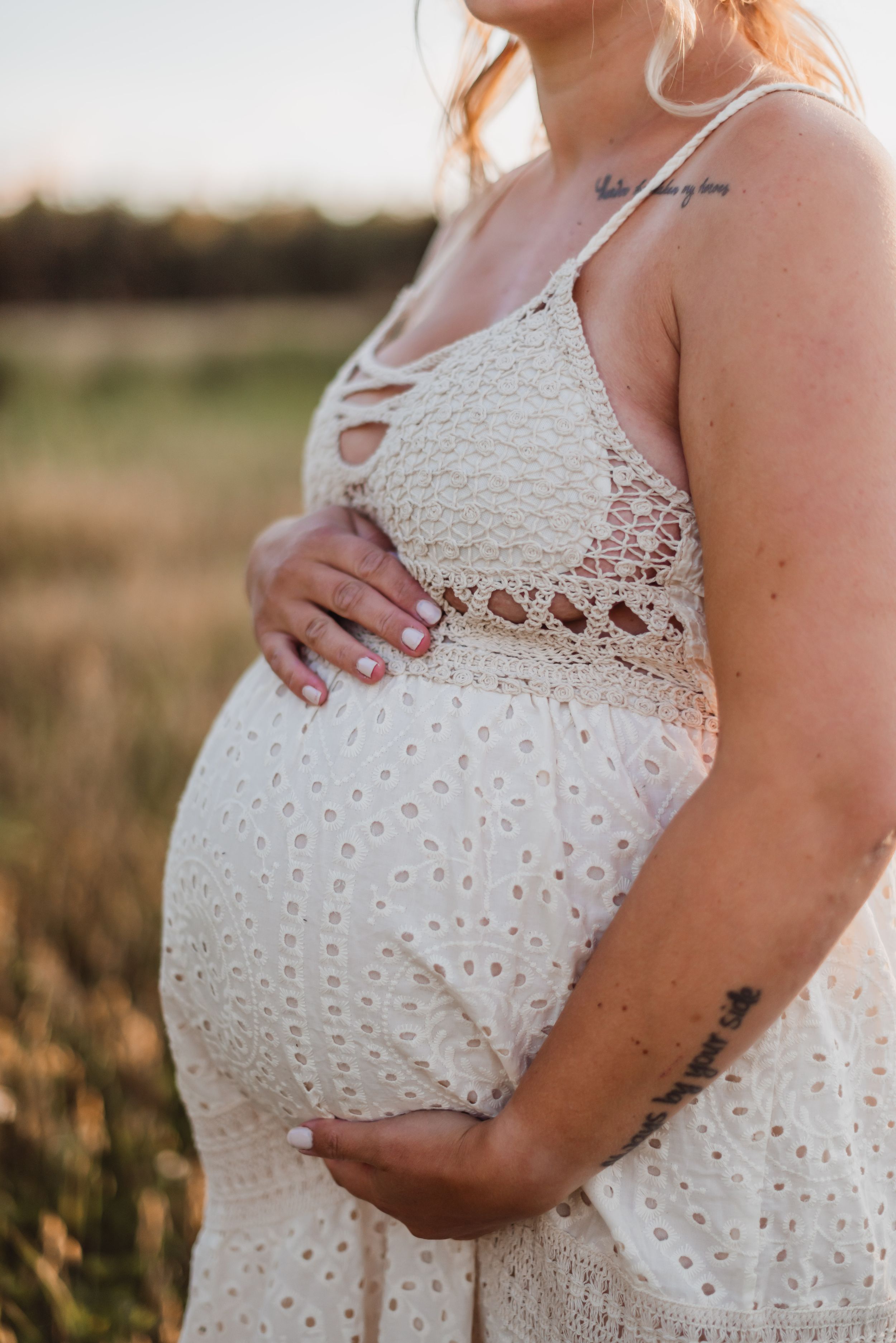Close-up maternity photo showing pregnant belly in white lace dress against golden sunset light in outdoor field setting.