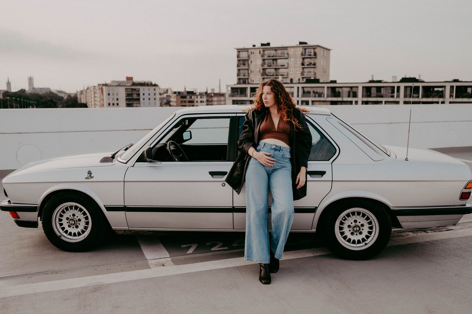 Classic white BMW E28 parked on rooftop with urban backdrop and retro fashion styling.