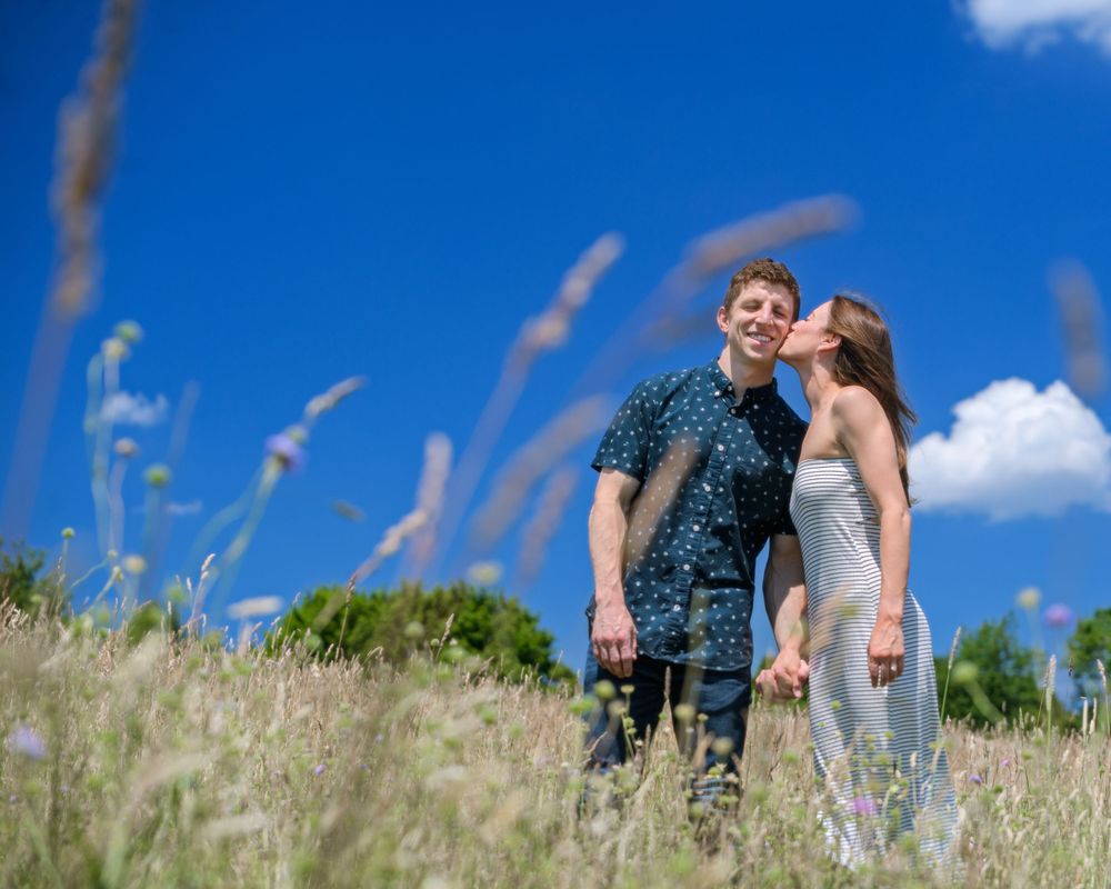 A couple standing in tall grass with blue sky and white clouds behind them.
