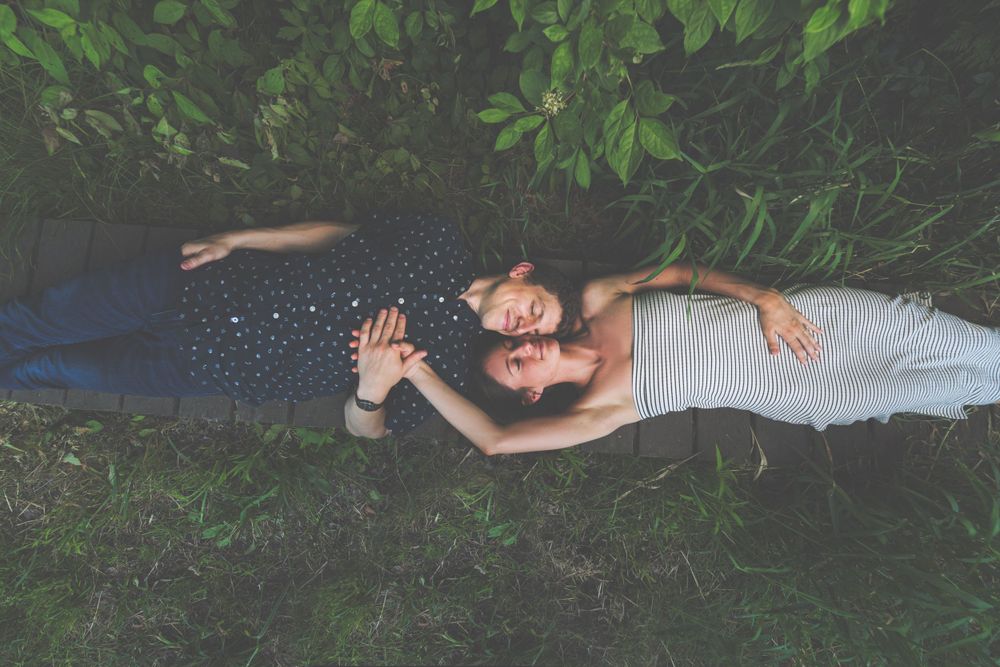 Two people lying on grass reaching their arms outward surrounded by green foliage.