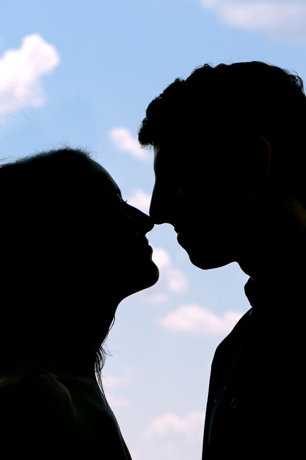 Silhouetted profiles about to kiss against a bright blue sky with fluffy white clouds.