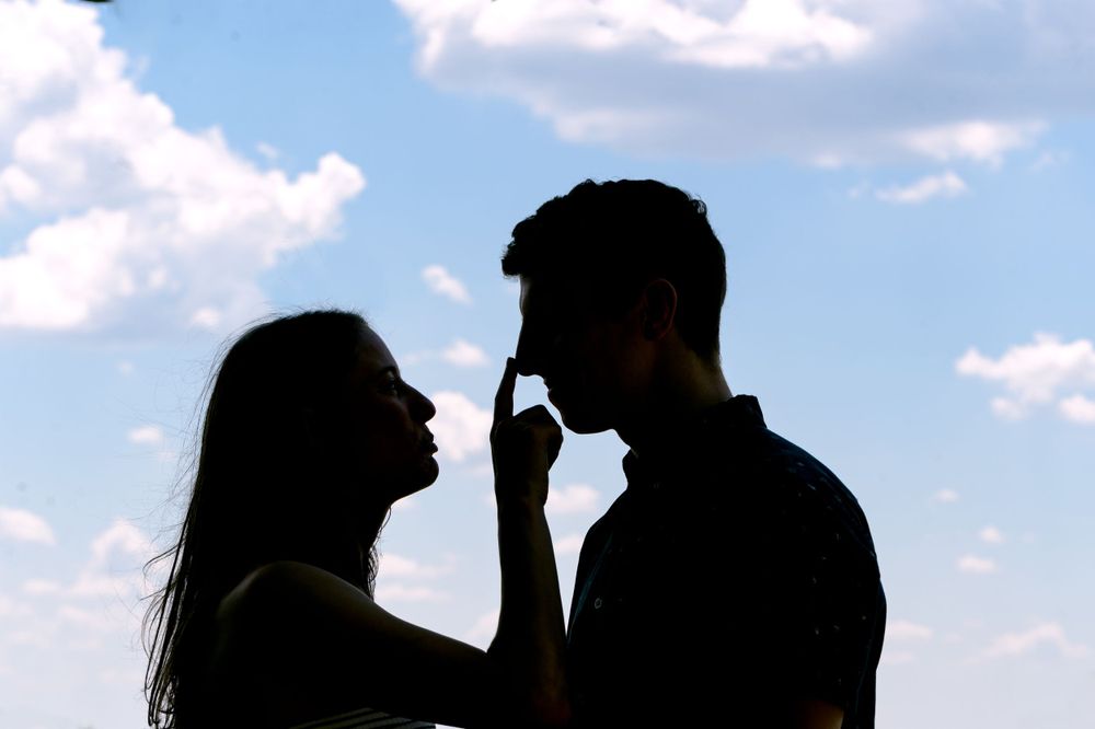 Silhouetted figures share an intimate moment against a bright blue sky with white clouds.