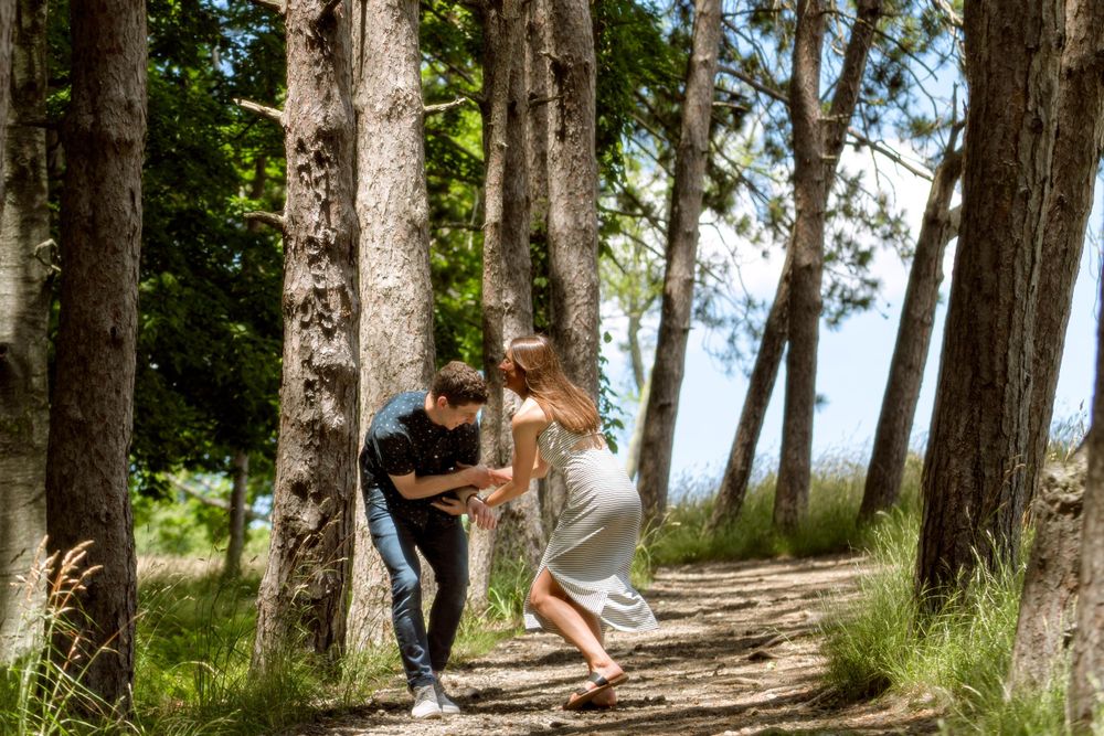 Two people playfully walking along a forest trail lined with tall pine trees.
