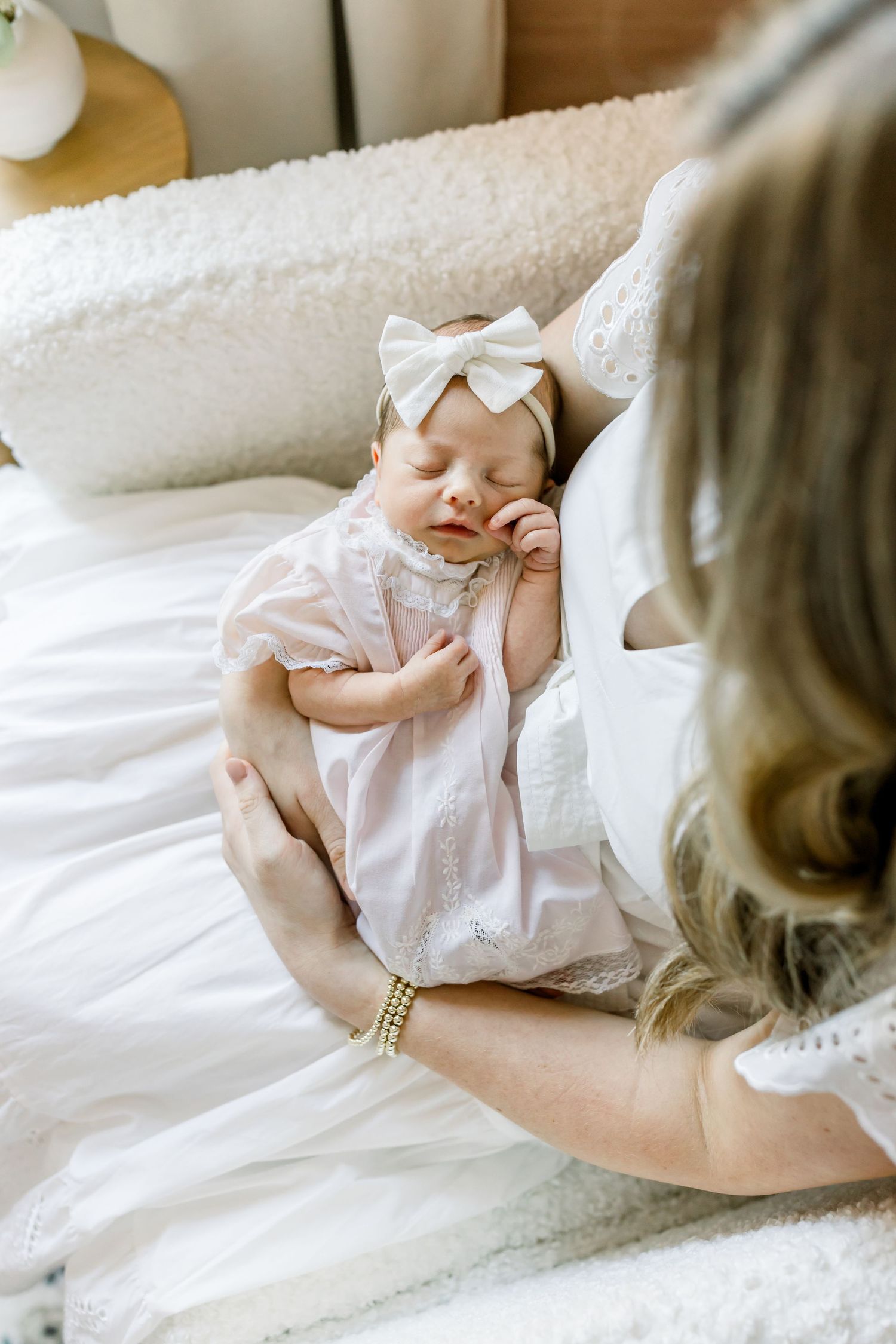 Sweet moment with sleeping baby in white dress and bow on white bedding.