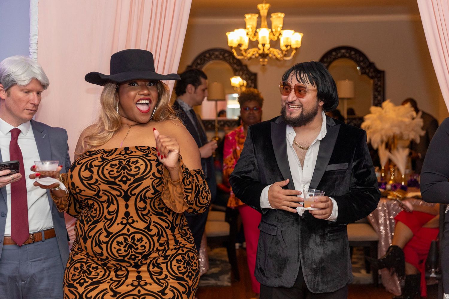A group of people laughing and socializing at a formal event with a chandelier and elegant decor in the background.