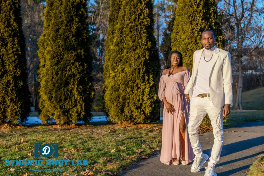 Couple in elegant pink dress and white suit pose in front of tall cypress trees.