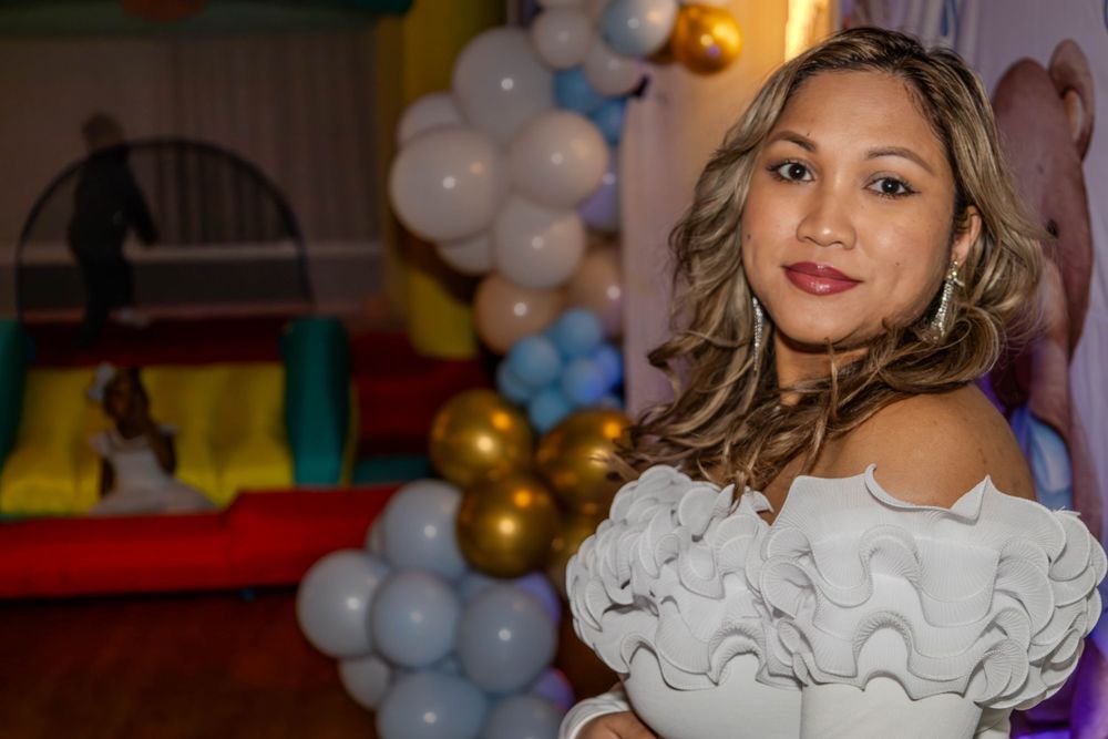 Elegant white off-shoulder dress at a baby shower with balloon decorations in the background.