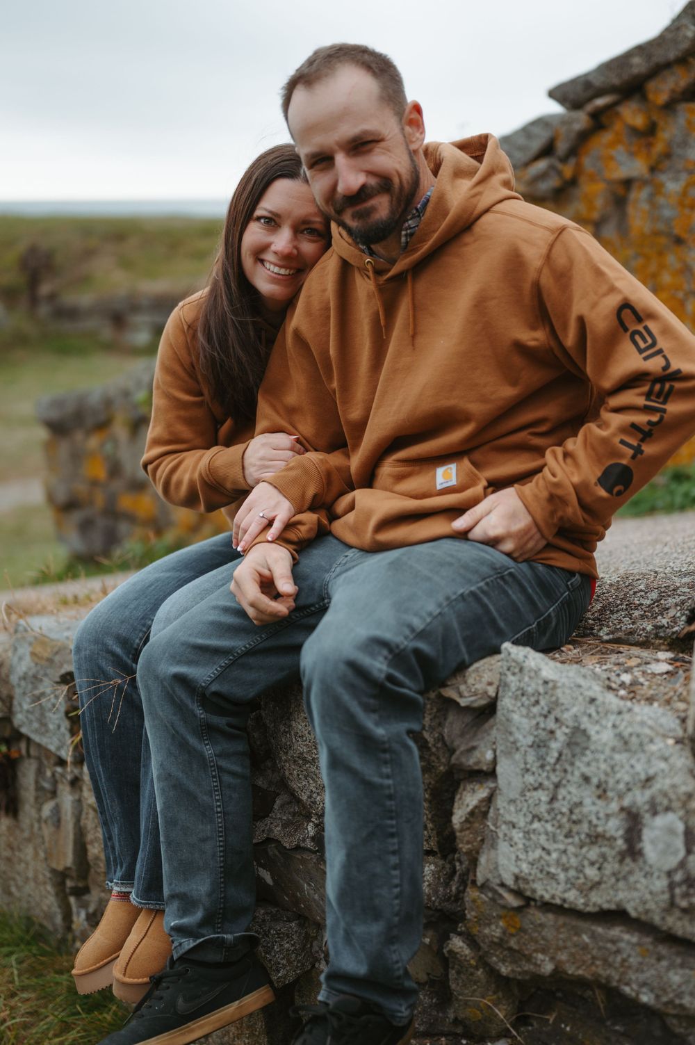 Two people sit together on an old stone wall wearing brown jackets on an autumn day.