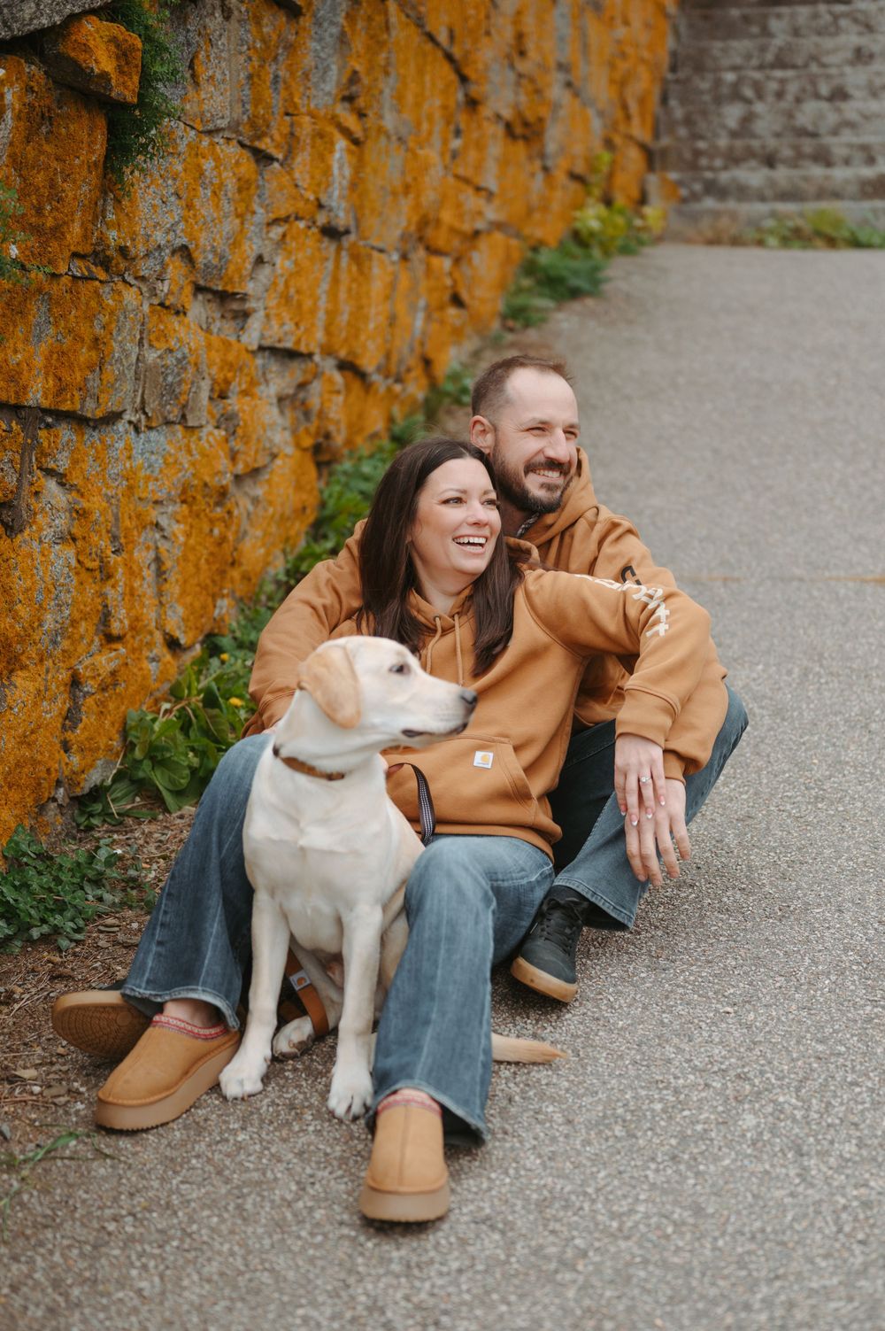 A couple and their white dog share joyful moments while sitting against an orange-tinted wall outdoors.