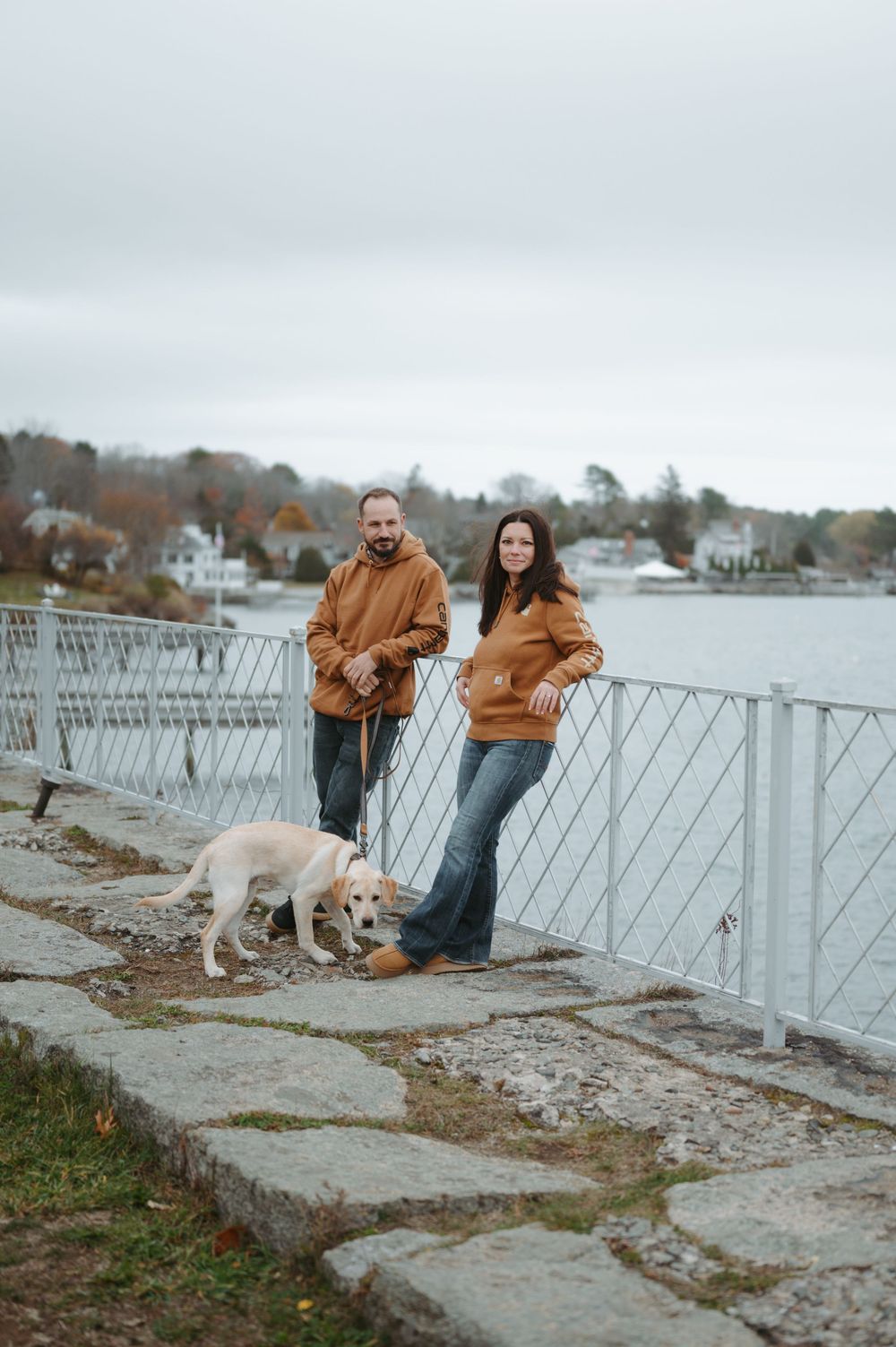 A couple in matching brown hoodies walks their white dog along a waterfront railing on an overcast day.