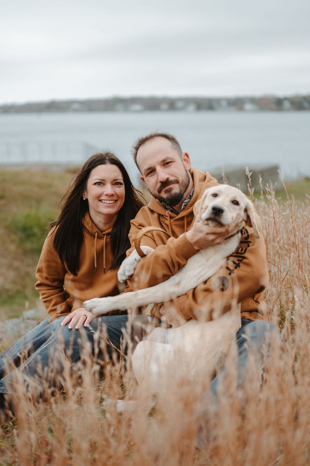 A couple in matching brown hoodies poses with their white dog in tall grass by the waterfront.