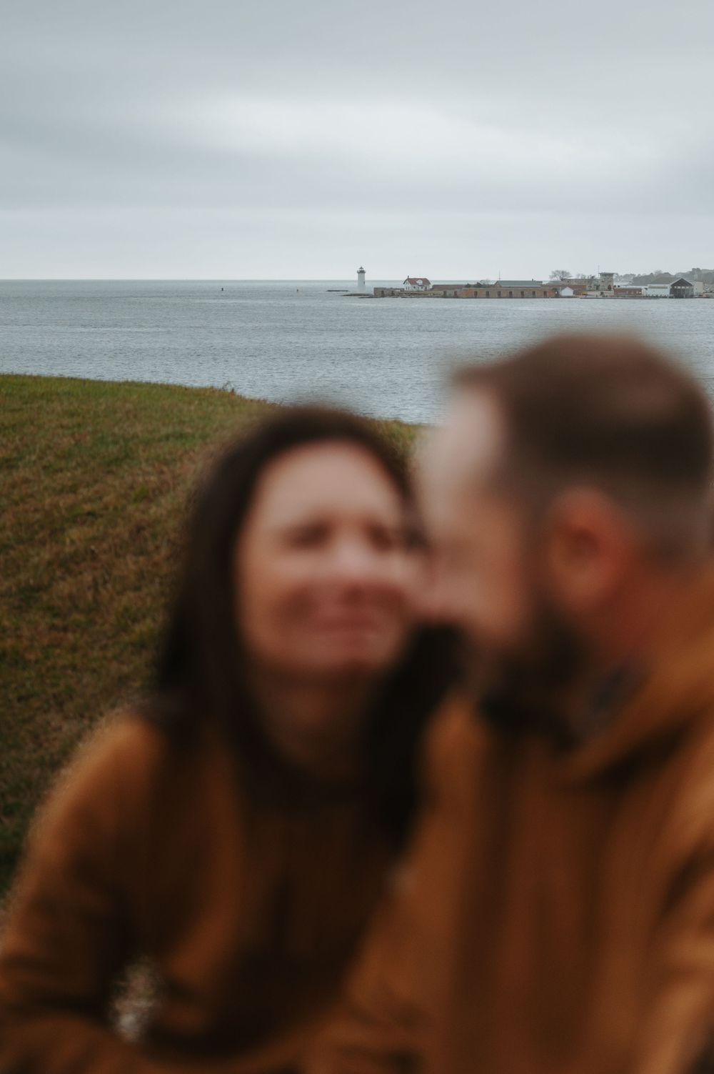A couple sits on a grassy hill overlooking a distant harbor on an overcast day.