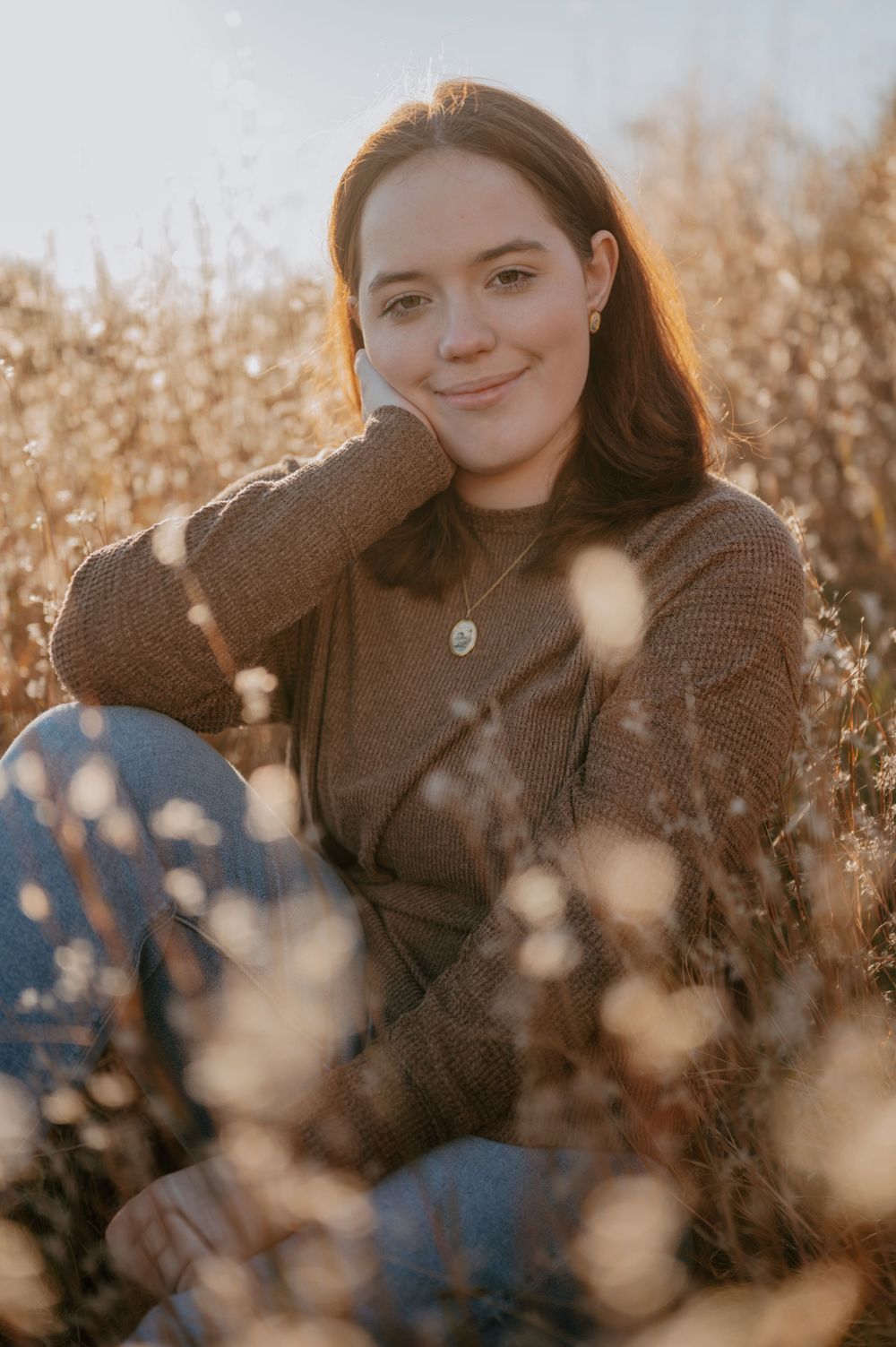 Person in brown sweater sitting outdoors during golden hour among dried plants.