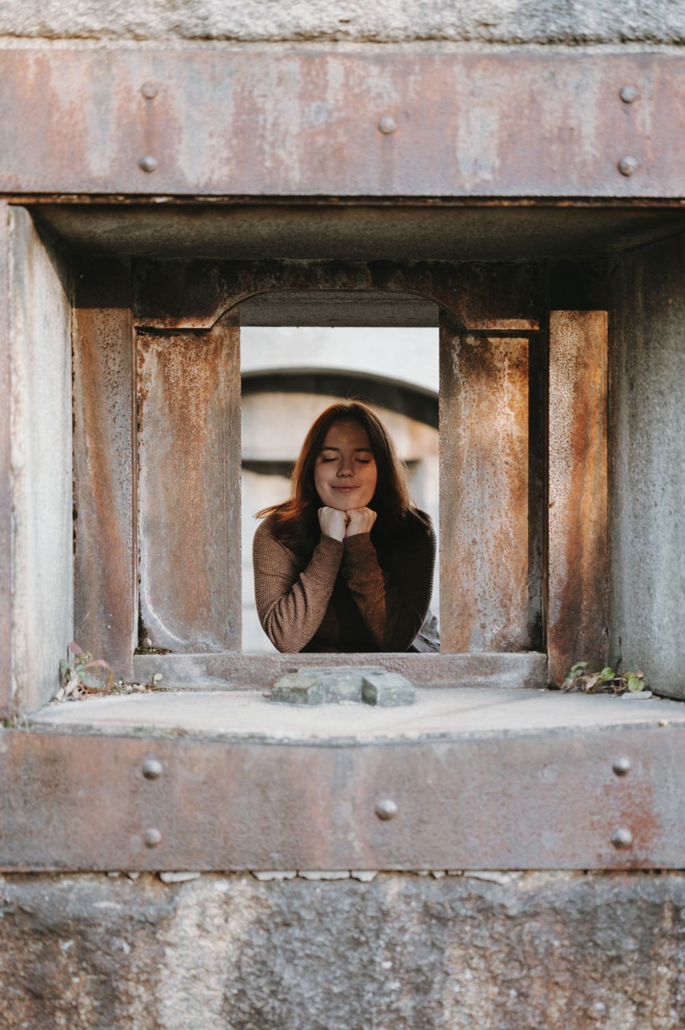 Person wearing brown sweater looking through rustic concrete window opening.
